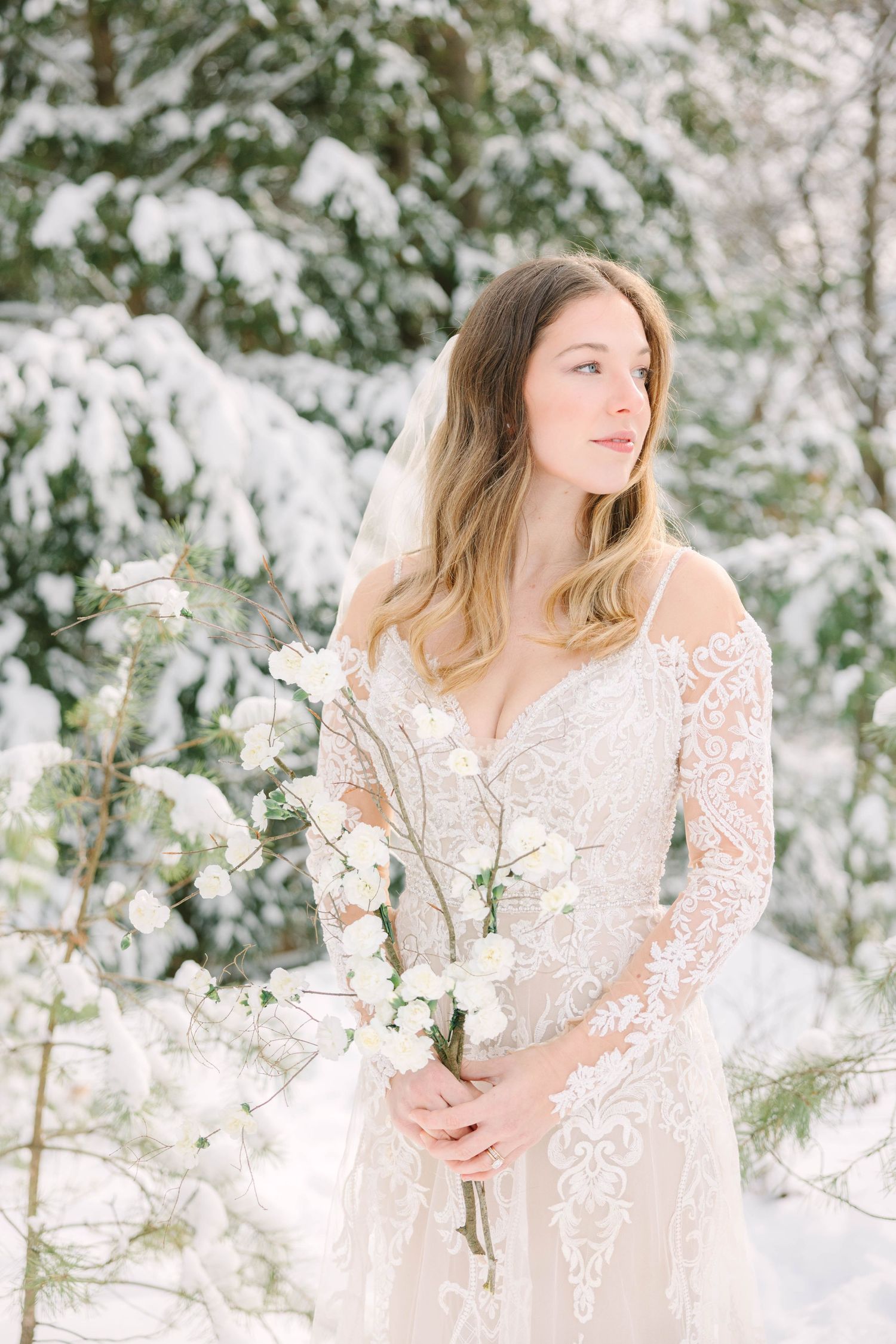 Bride in lace long-sleeve wedding gown holds white floral bouquet among snow-covered evergreen trees.