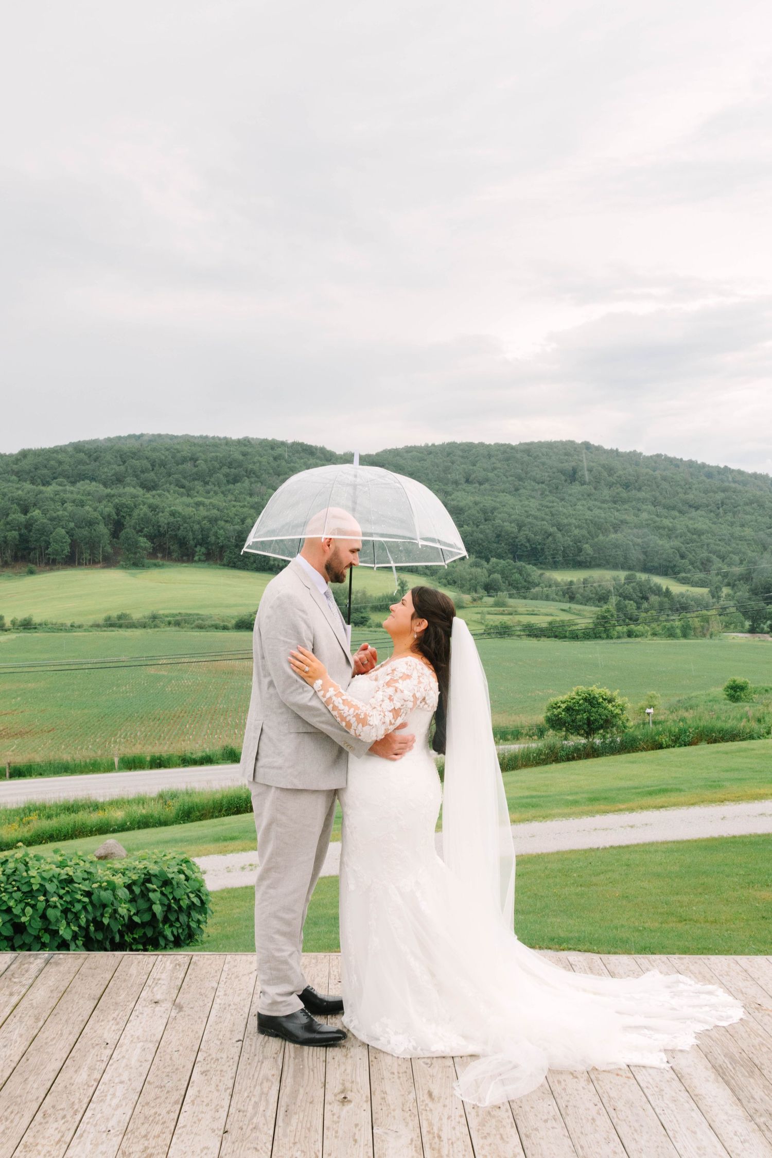 Bride and groom share a romantic moment under a clear umbrella on a wooden deck with lush green hills behind them.