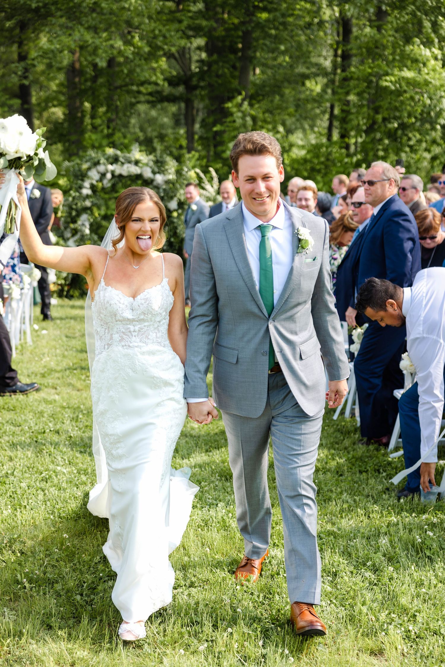 Joyful newlyweds walk back down grassy aisle hand in hand after outdoor wedding ceremony.