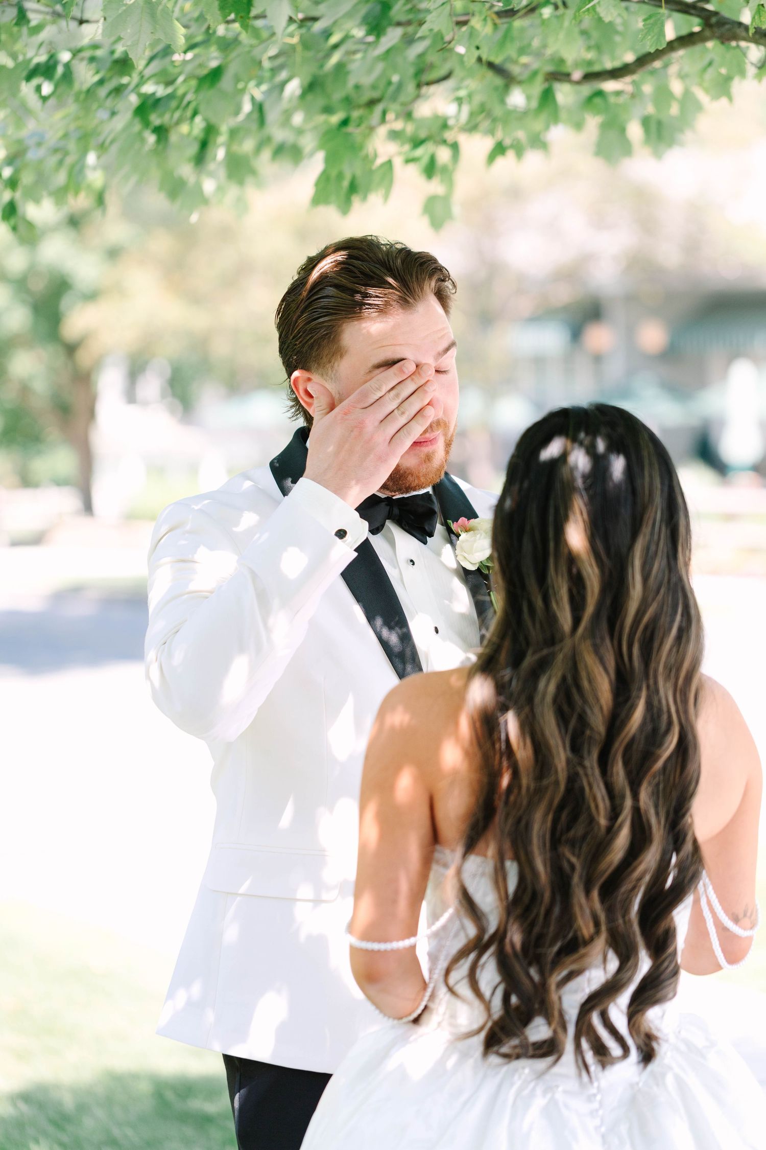 Groom in white tuxedo covering face with hand in emotional reaction during wedding first look.