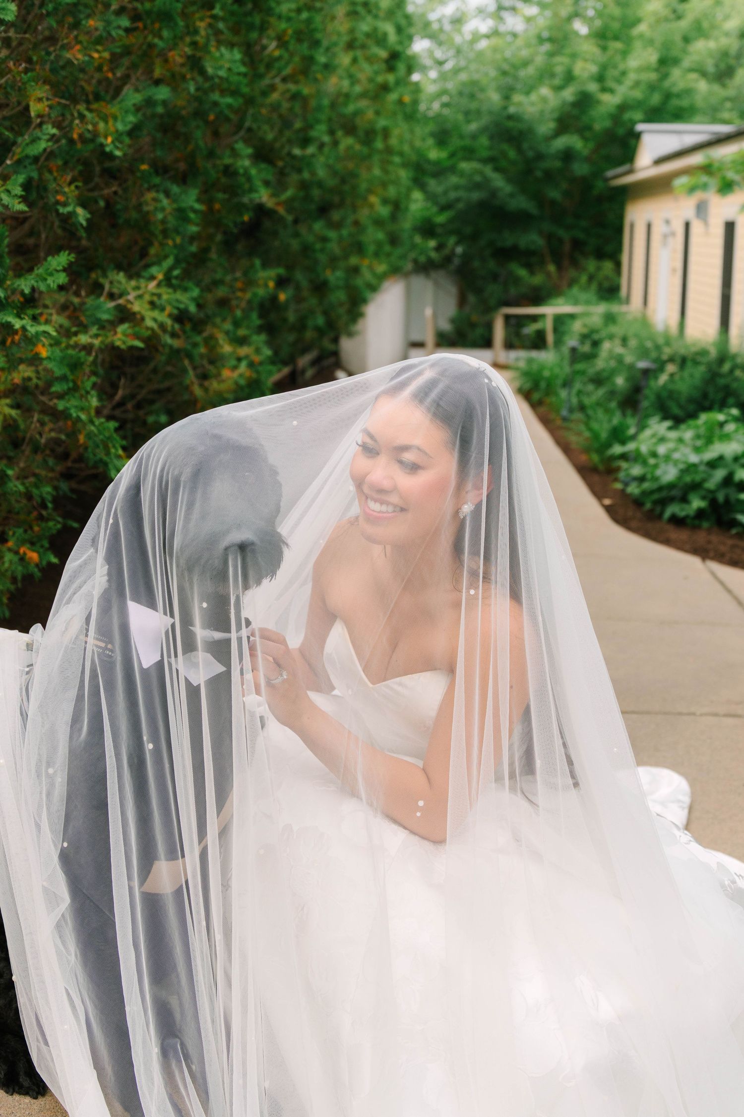 Smiling bride covered by flowing white veil, wearing strapless wedding gown in a lush garden setting.