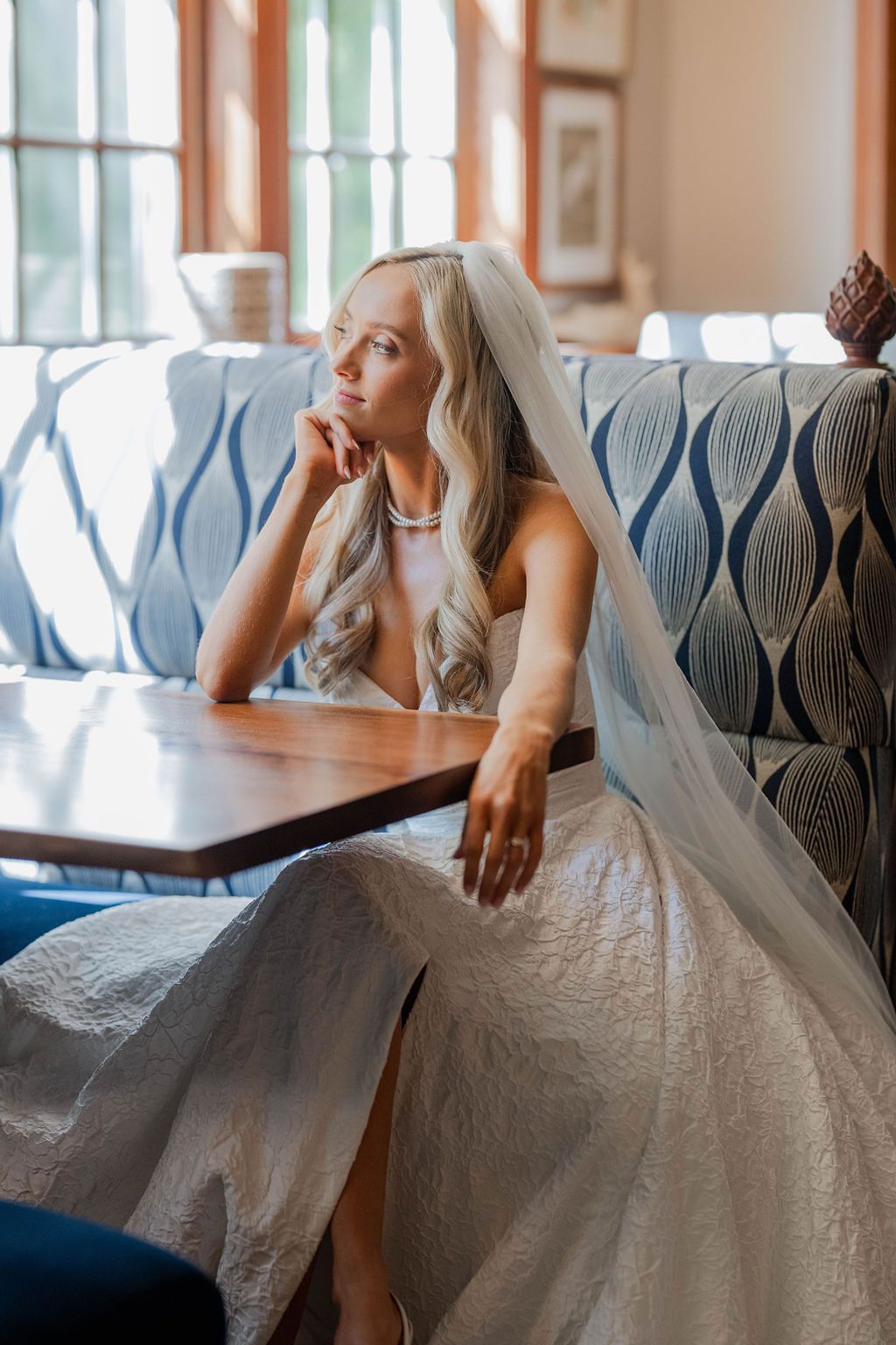 Bride in white lace gown and veil sits pensively at a wooden table on a blue patterned sofa.