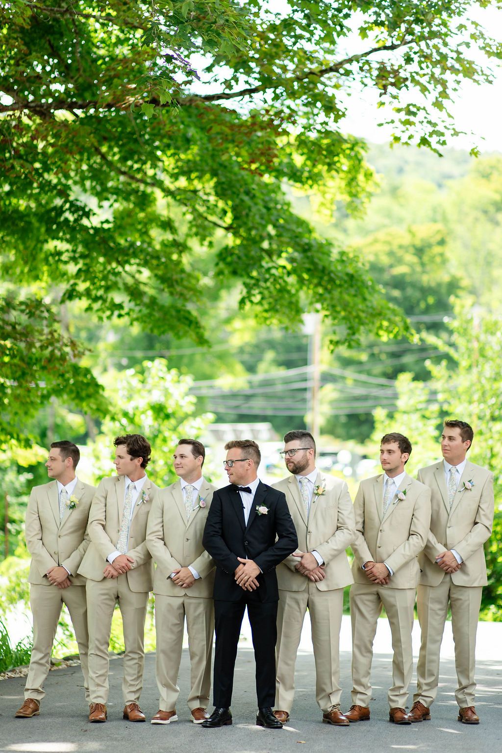 Groom in black tuxedo stands with groomsmen in tan suits outdoors under green trees at wedding.