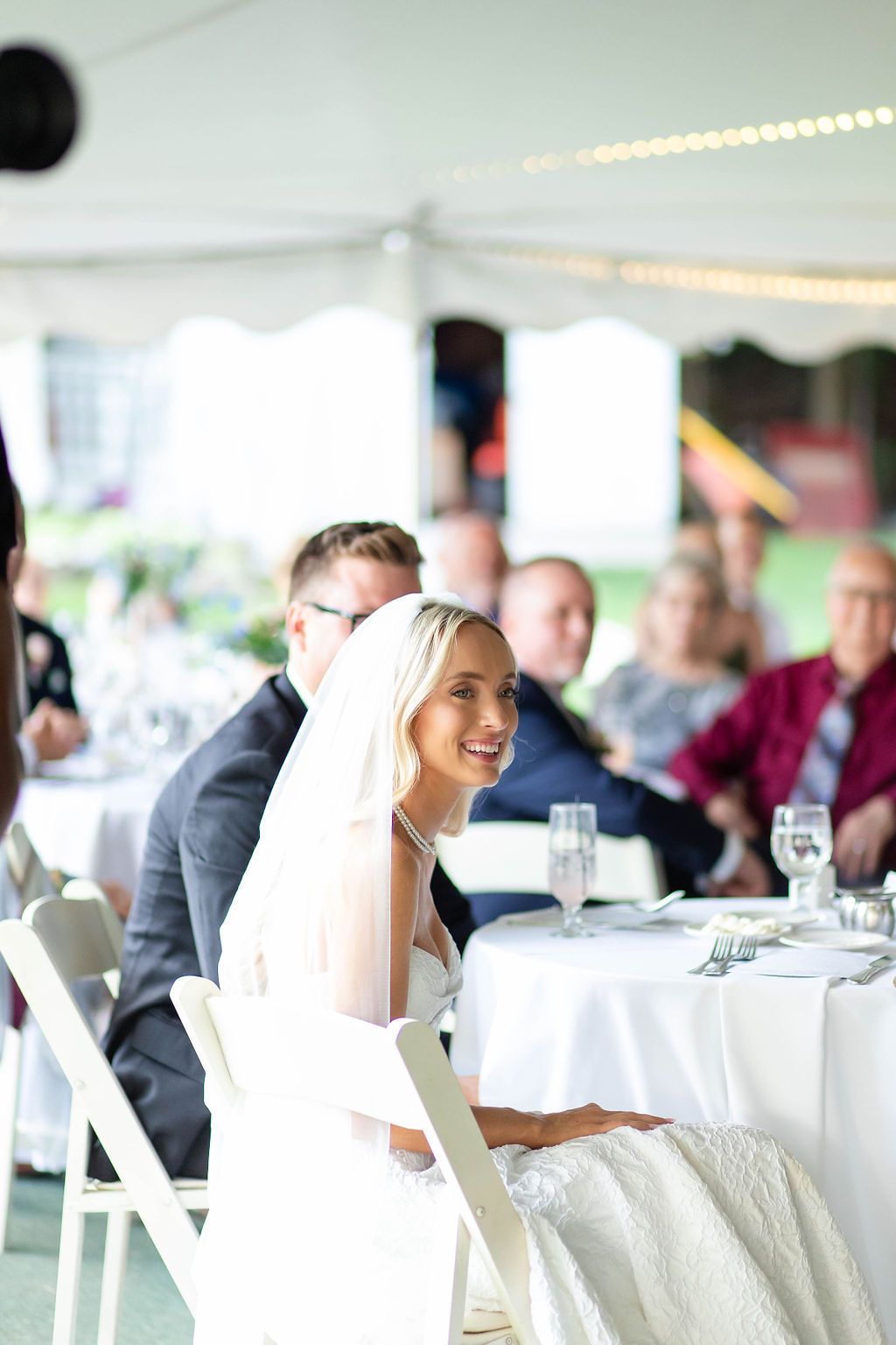Smiling bride in white gown and veil seated at head table during outdoor wedding reception under white tent.