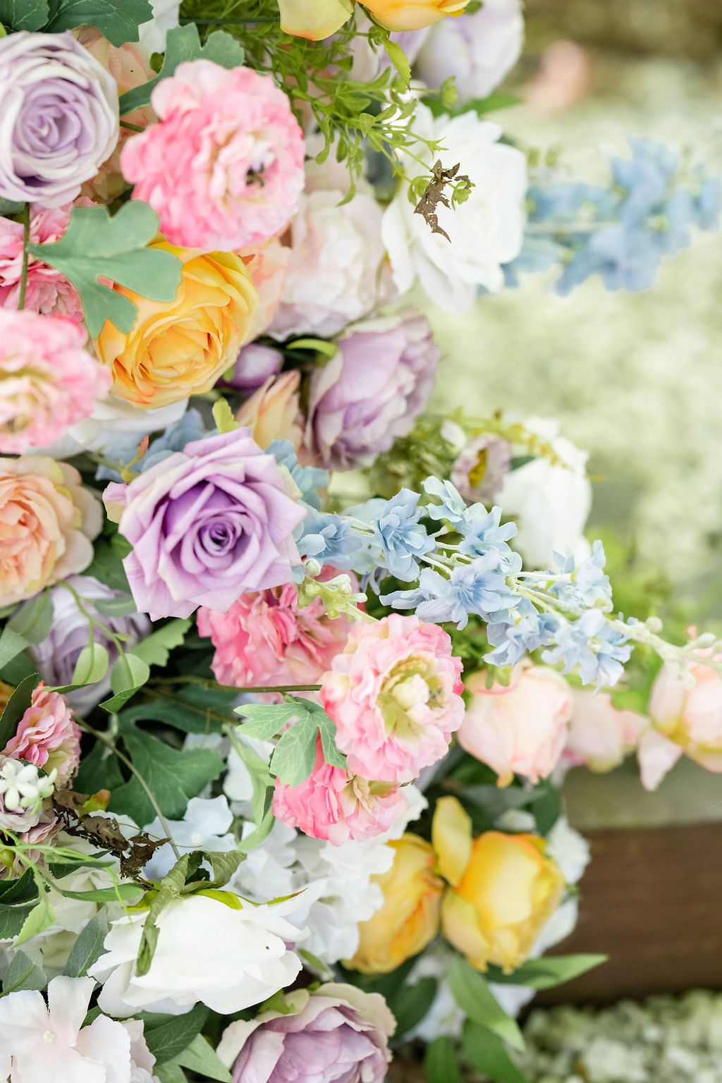 Colorful wedding floral arrangement featuring pastel roses in pink, purple, yellow, and white with blue accent flowers.