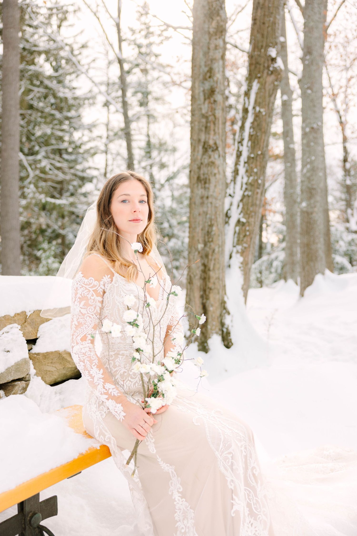 Bride in lace wedding dress and veil sits on snowy bench holding white flowers in winter forest.