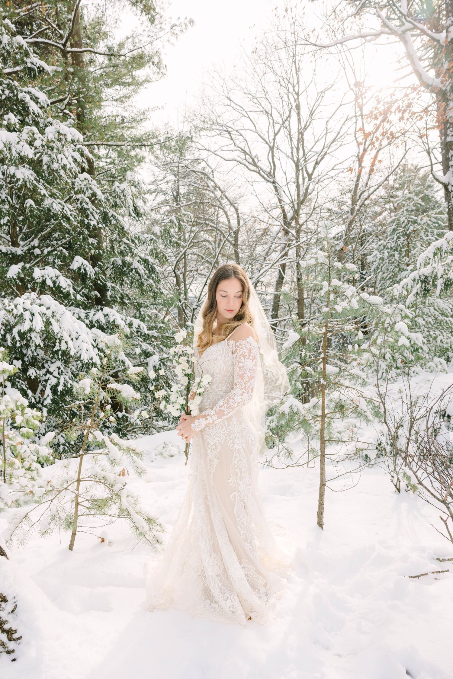 Bride in lace gown standing in snowy winter forest surrounded by snow-covered pine trees.