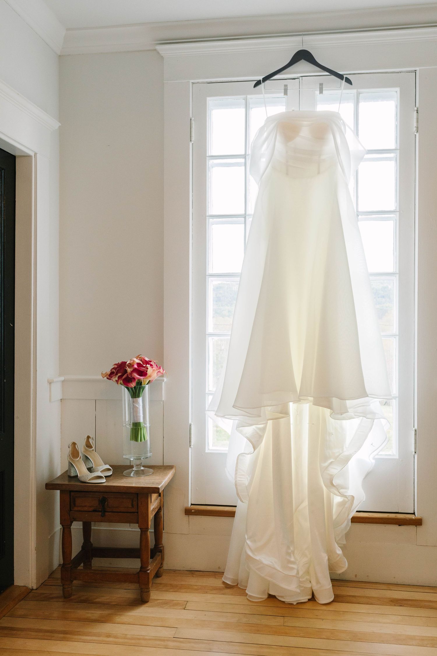 White wedding gown hanging on hanger in front of bright window with bridal shoes and pink flowers on wooden side table.