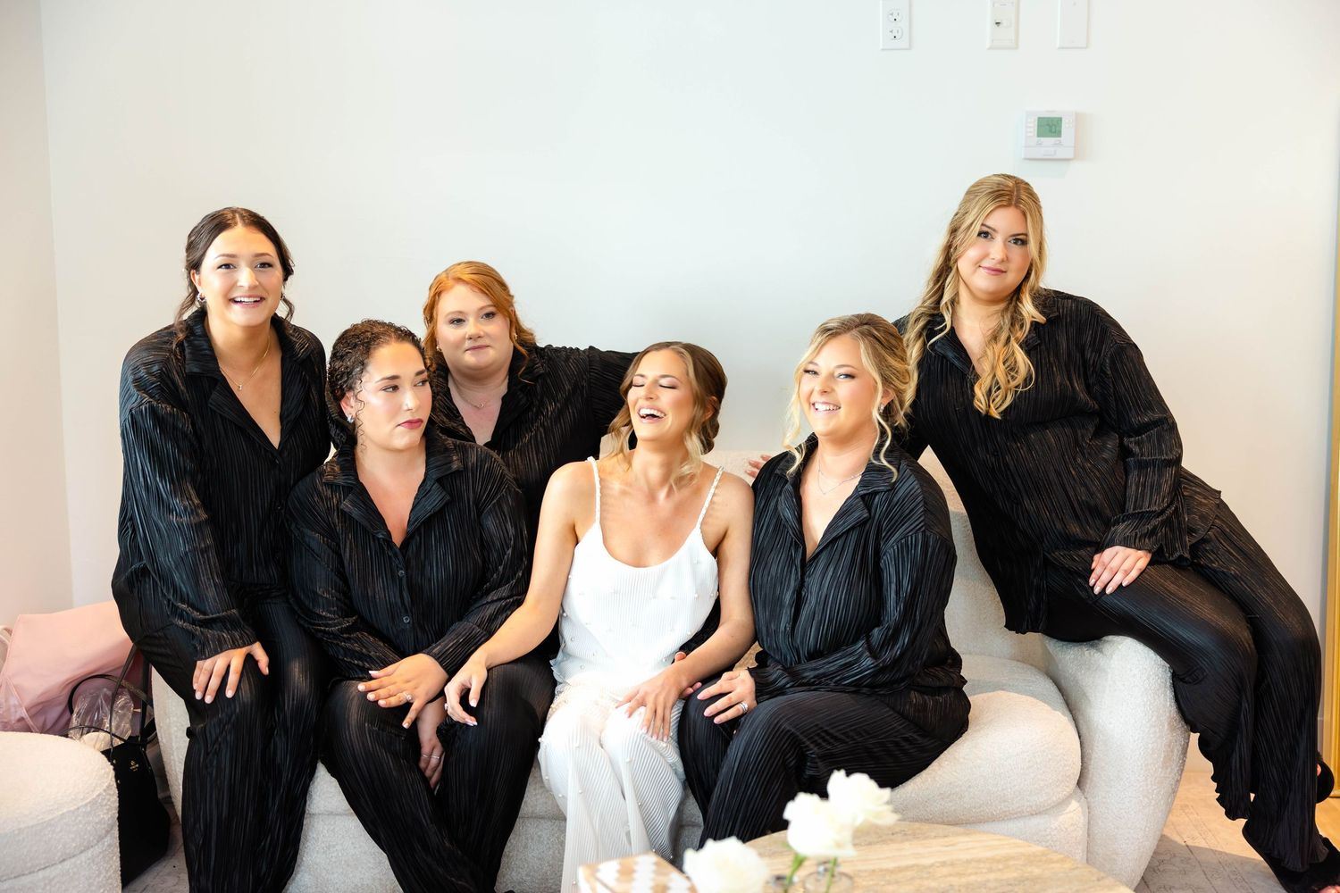 Bride in white dress sits with bridesmaids wearing black satin pajamas in bright room.