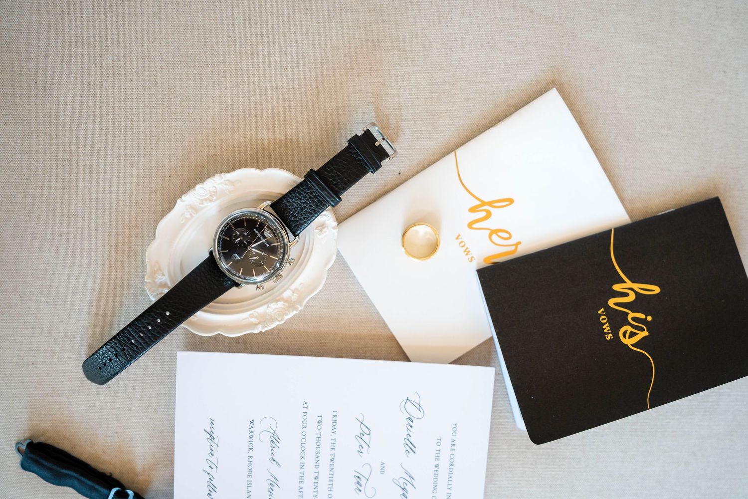 Wedding flat lay featuring a black watch, gold ring, invitation cards, and a black and gold groom booklet on a neutral background.