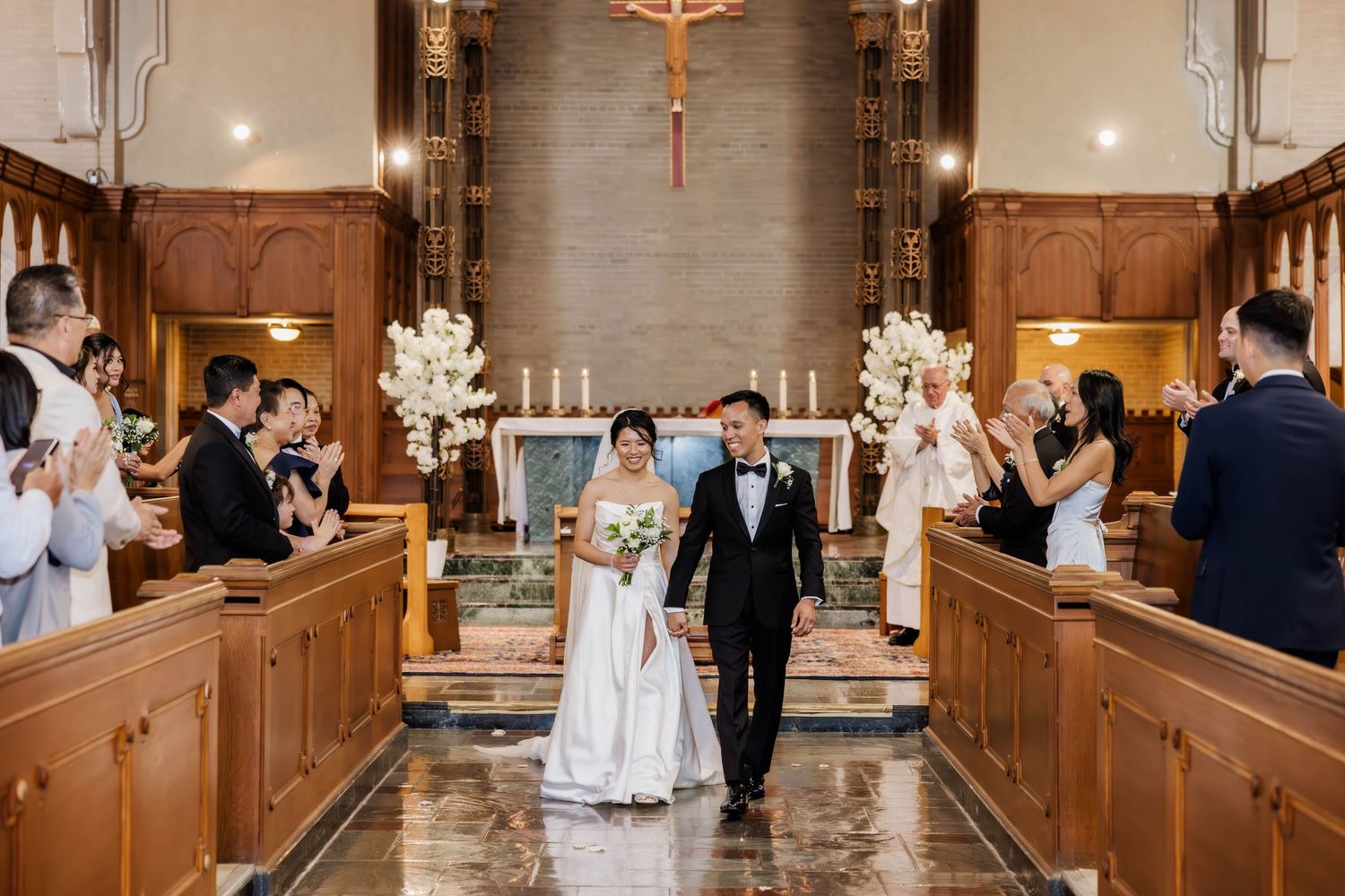 Bride and groom walking down church aisle after ceremony as guests applaud in elegant chapel.