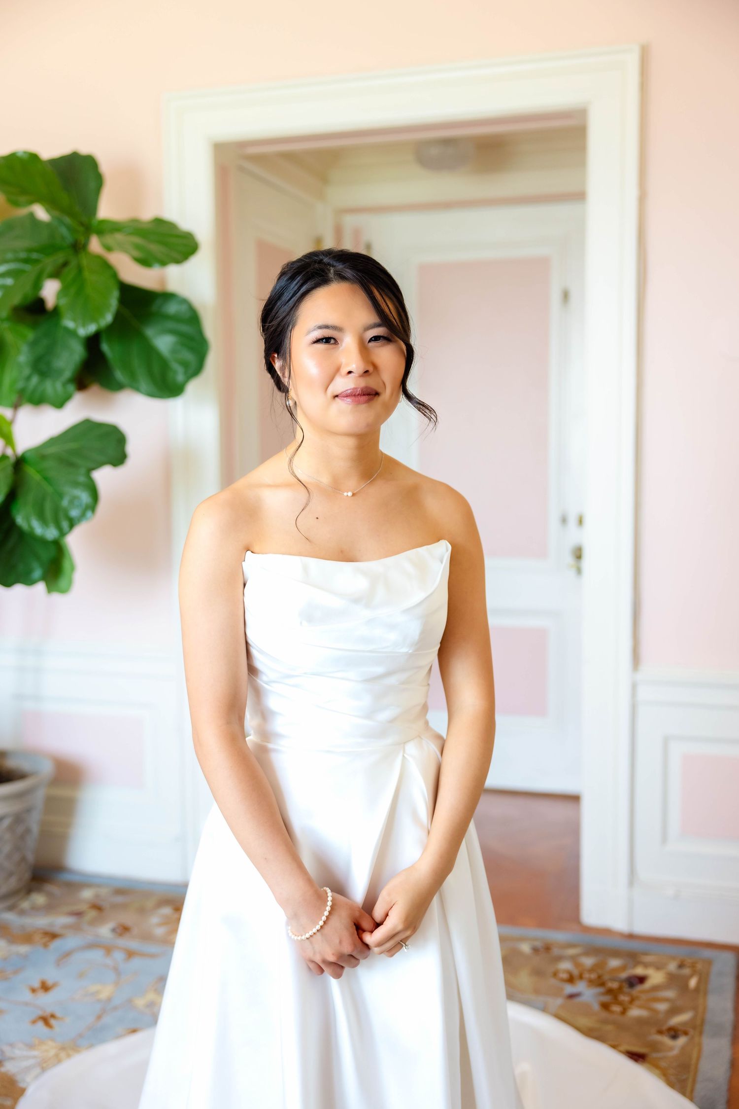 Bride in strapless white satin gown stands elegantly in a pink-walled room with a fiddle leaf fig plant.