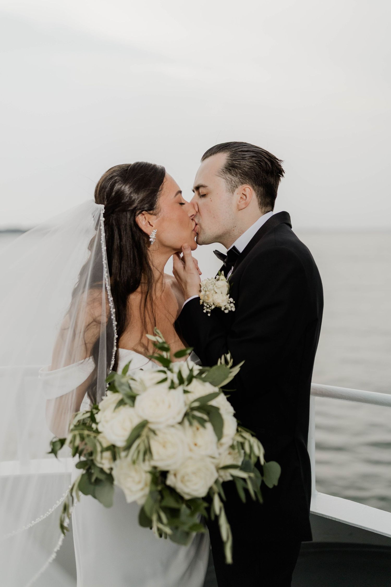 Bride and groom share romantic kiss on boat deck with white rose bouquet and bridal veil.