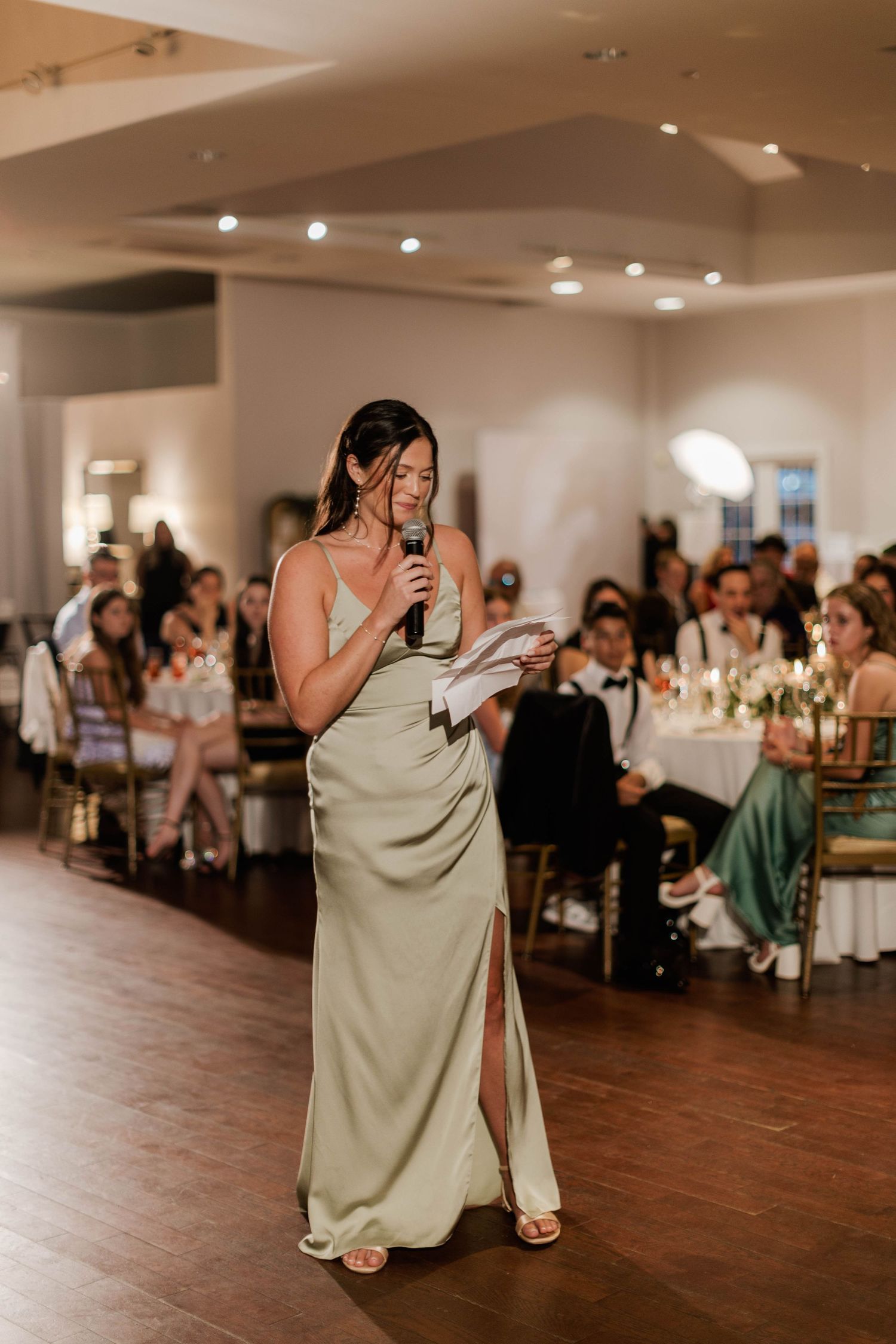 Woman in sage green satin gown reads a wedding toast while holding a microphone on the dance floor.