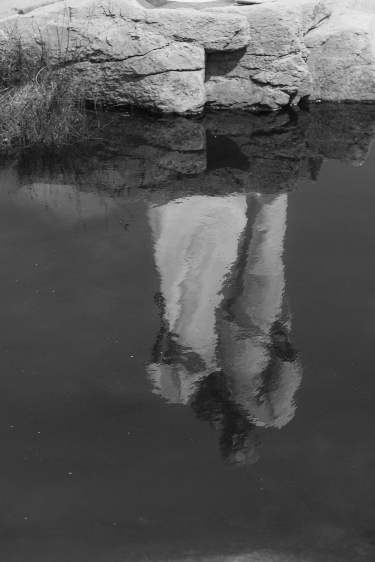 Black and white photo of a person's reflection in still water beside large rocks.