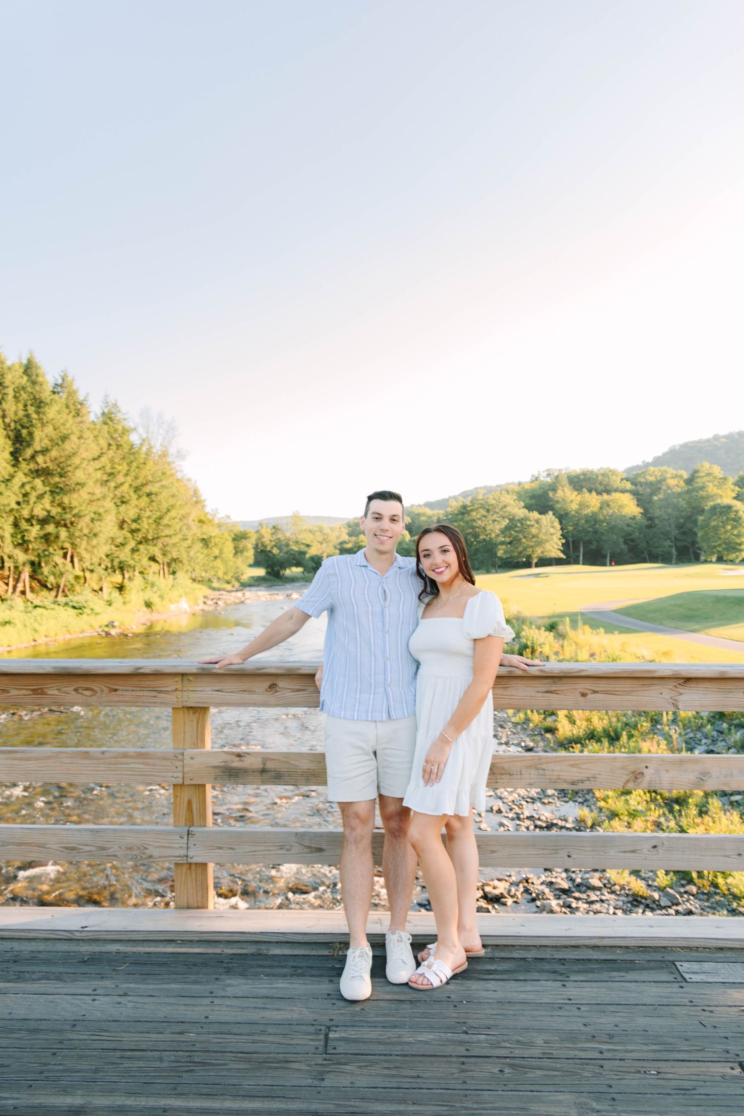 A couple smiles together on a wooden bridge overlooking a river and green hills on a sunny day.