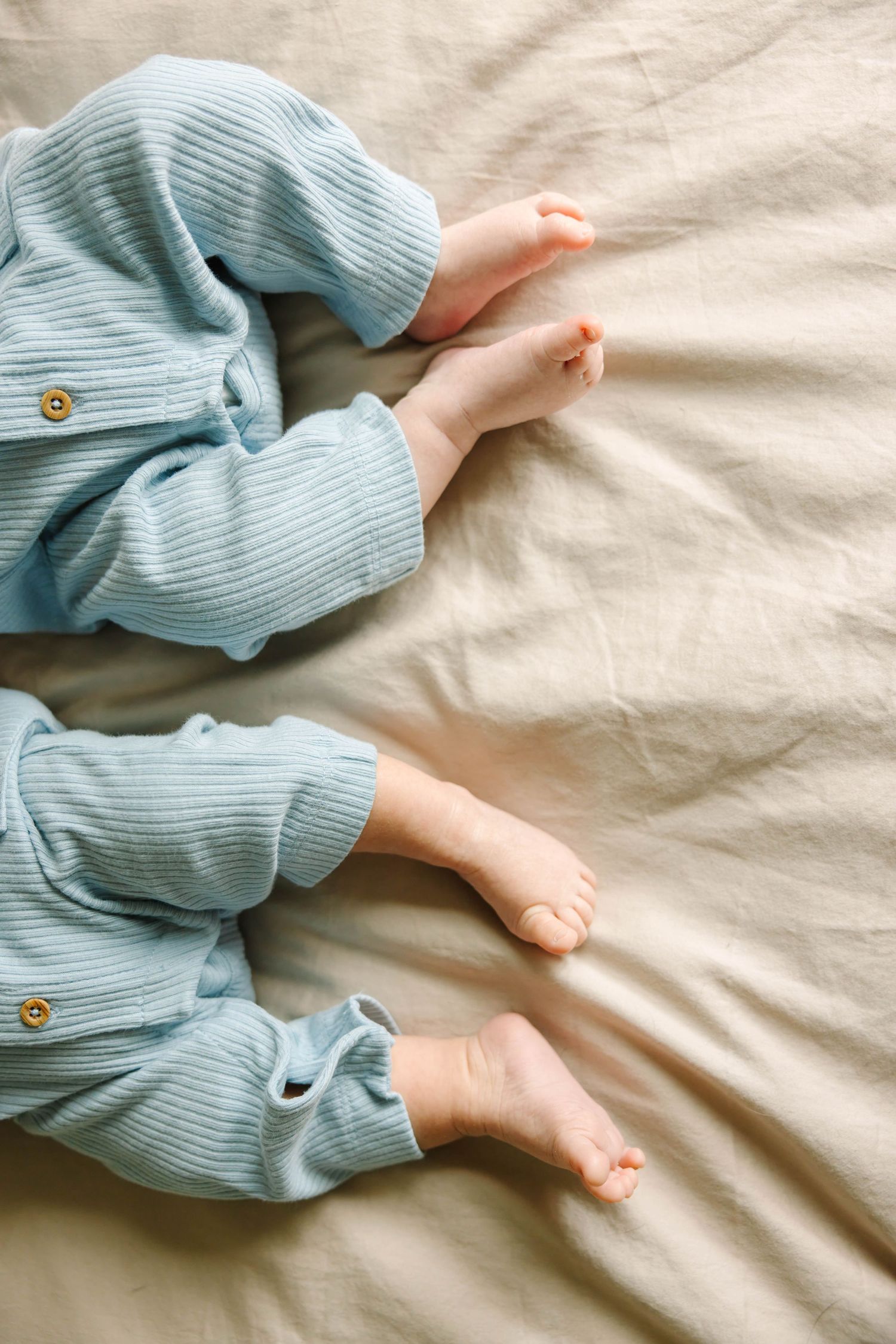 Close-up of two newborn twins' bare feet in matching blue ribbed rompers on beige linen.