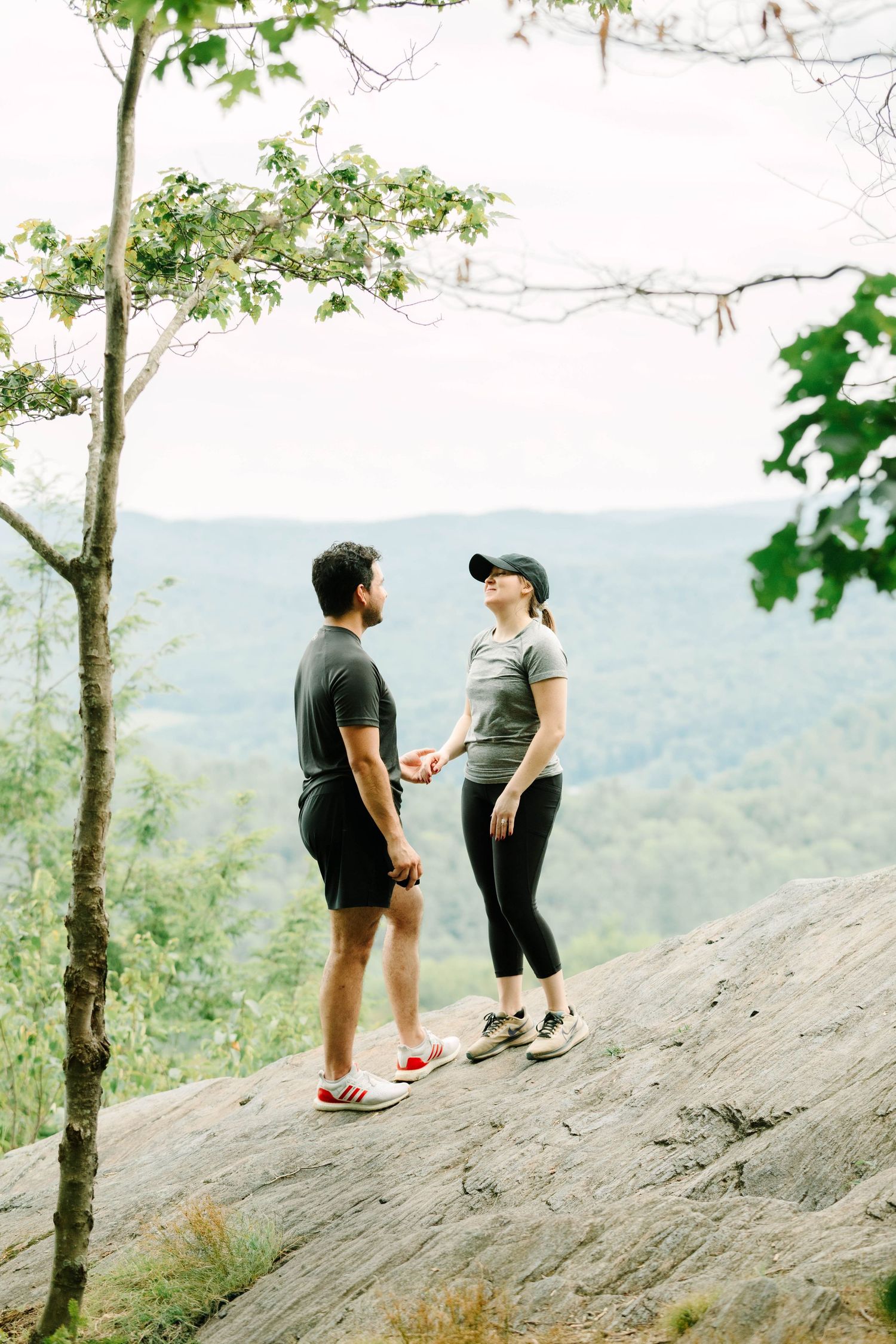 Couple holding hands on a rocky summit surrounded by trees with rolling green hills in background.