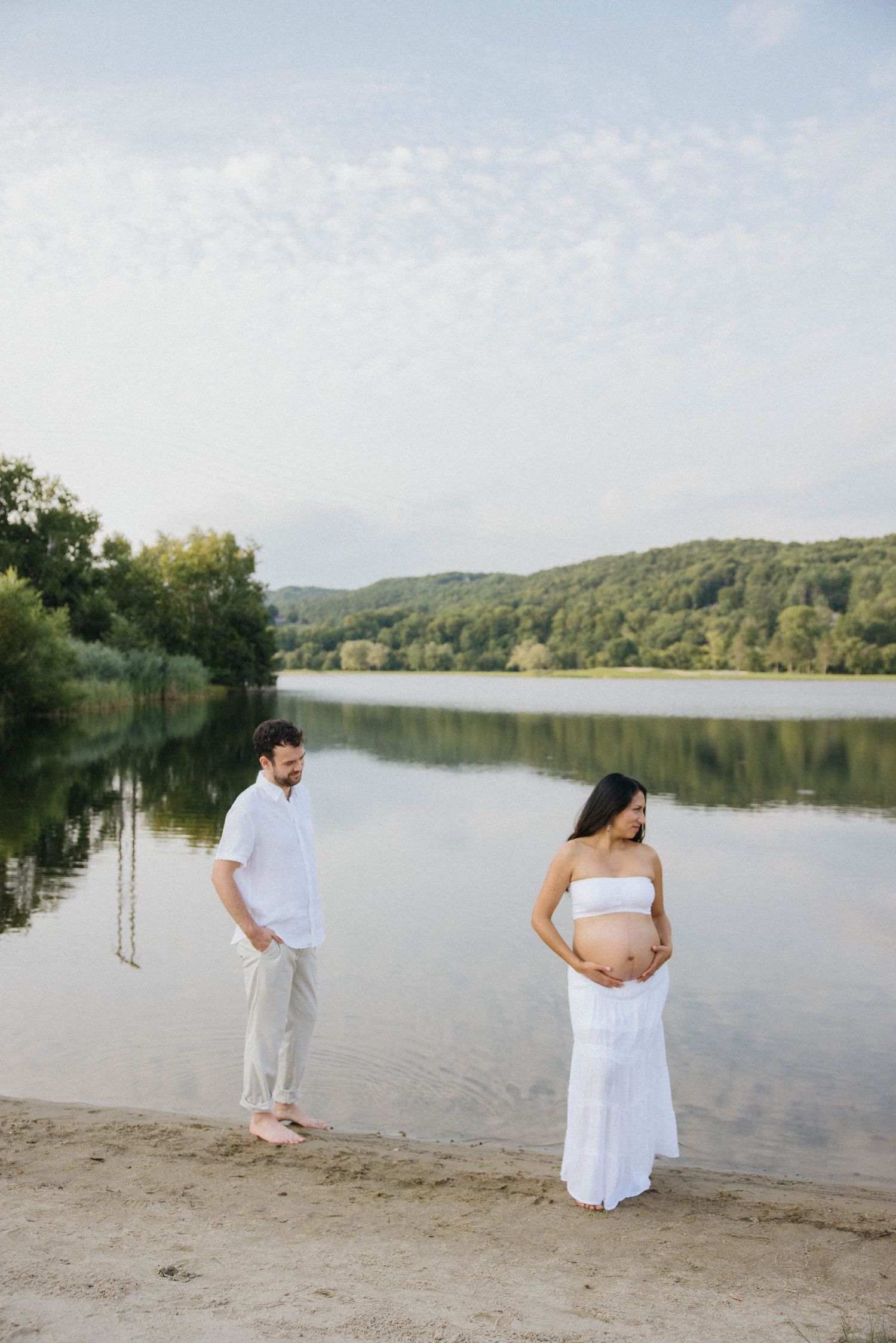 Pregnant woman in white dress holds belly by lake, man in white linen stands nearby on sandy shore.