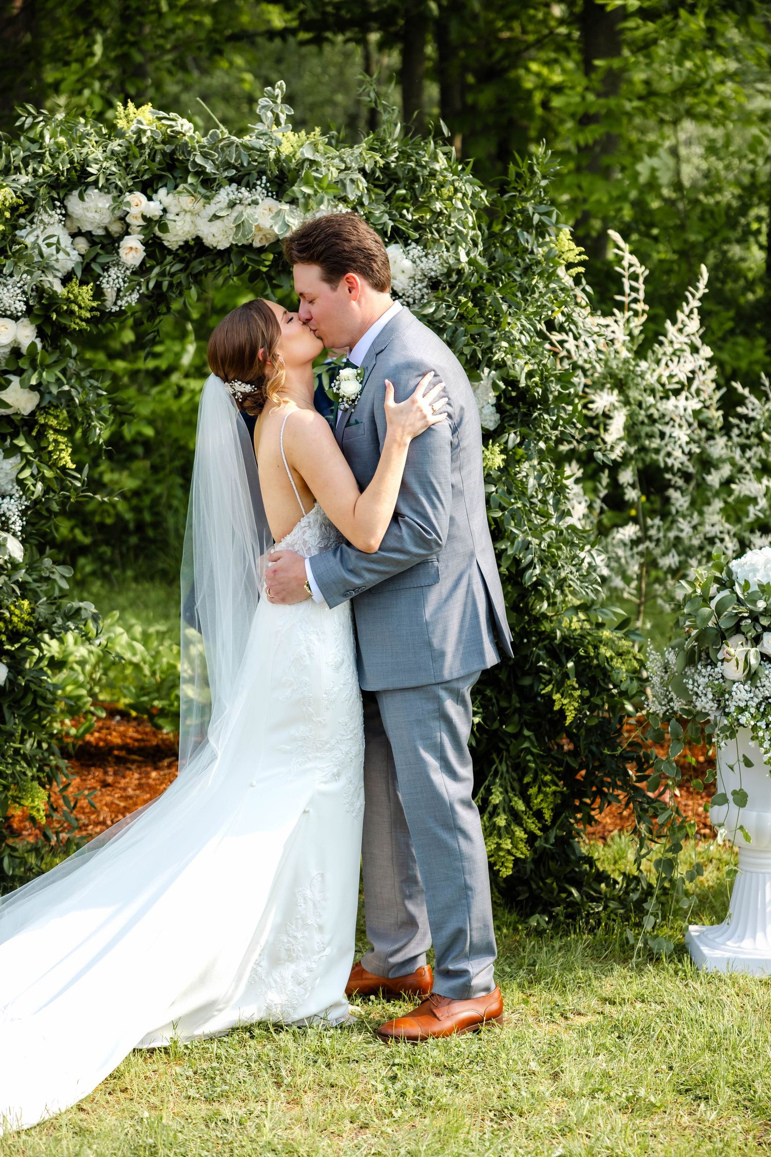 Newlyweds share first kiss under lush greenery and white floral arch at outdoor wedding ceremony.