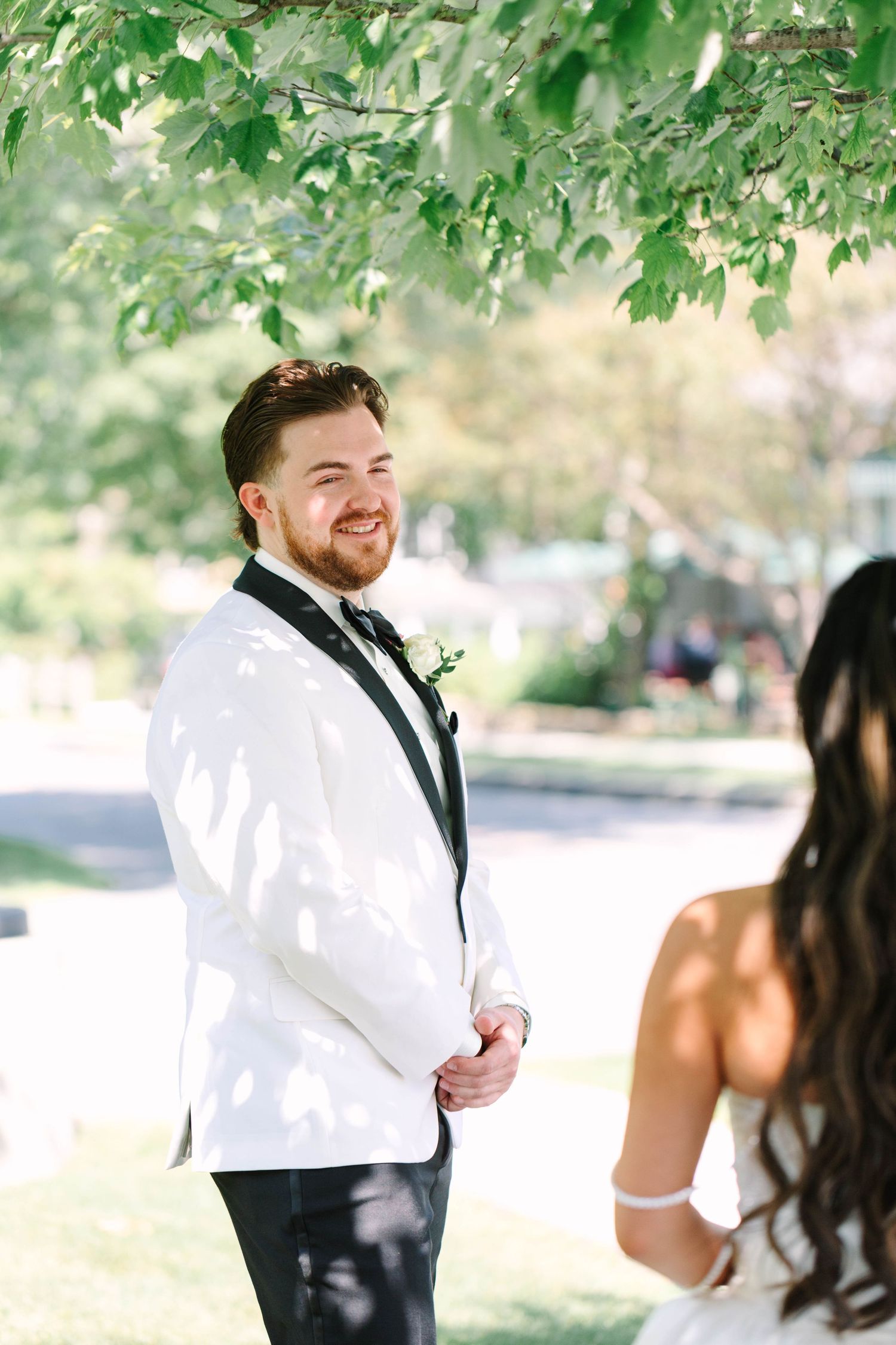 Emotional groom in white tuxedo smiling at bride during outdoor first look wedding moment.