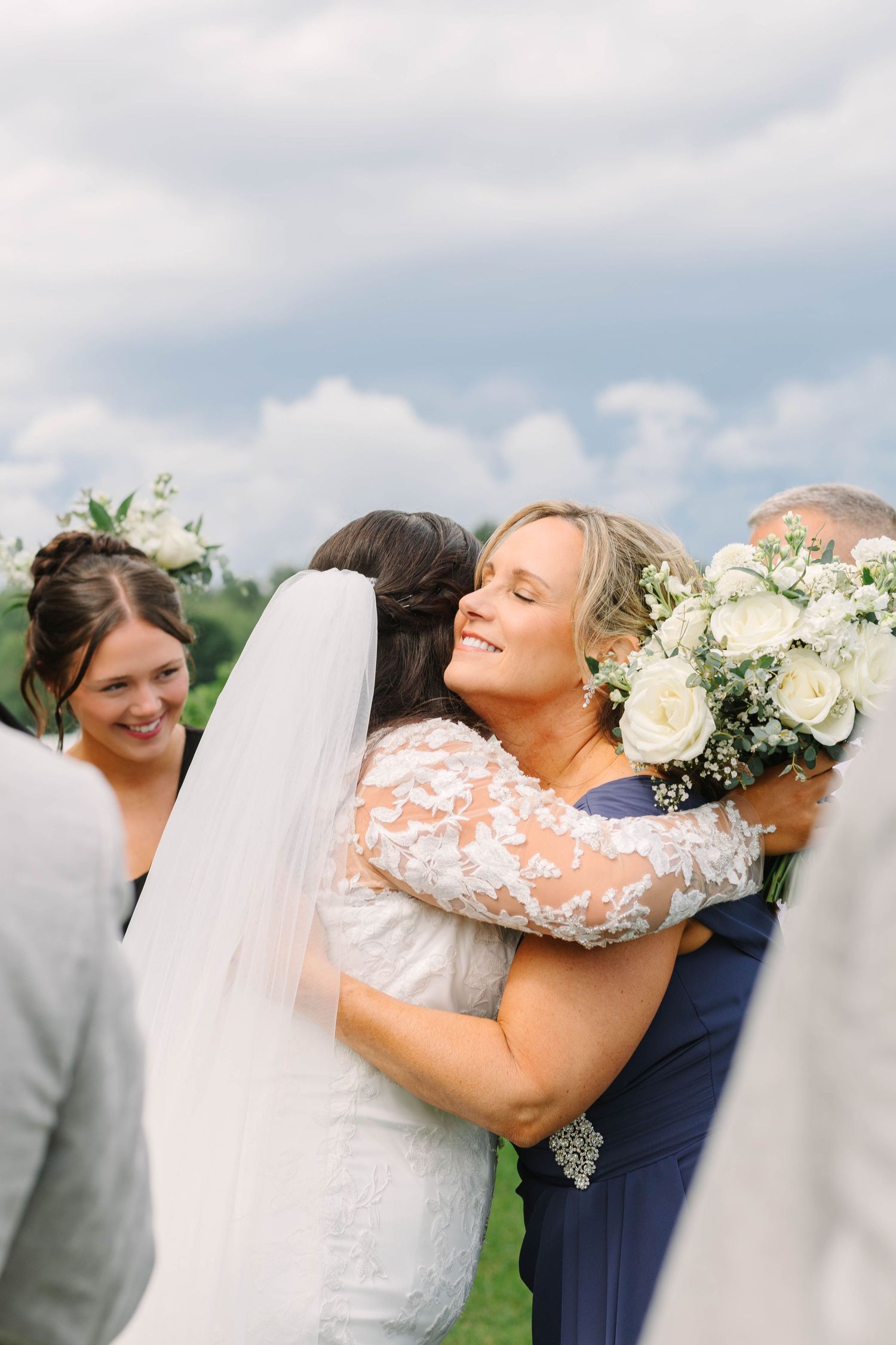 Bride shares a warm embrace with a smiling woman in navy blue during outdoor wedding ceremony.