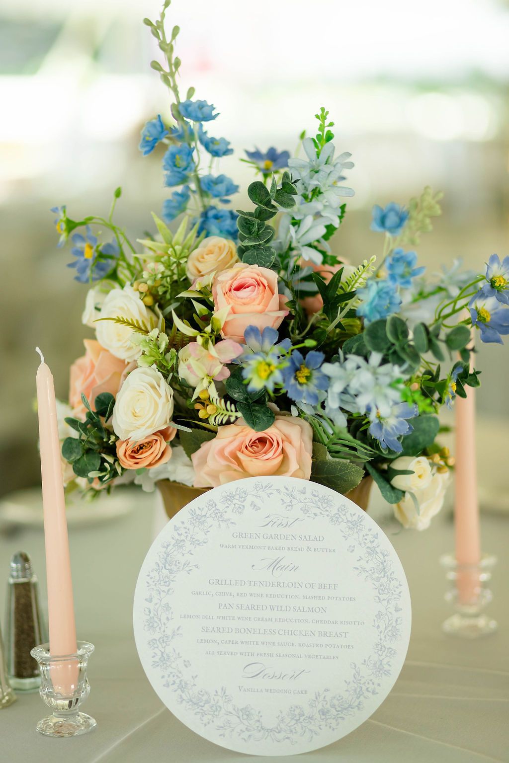 Round wedding menu card beside pink candles and blue and peach floral centerpiece.