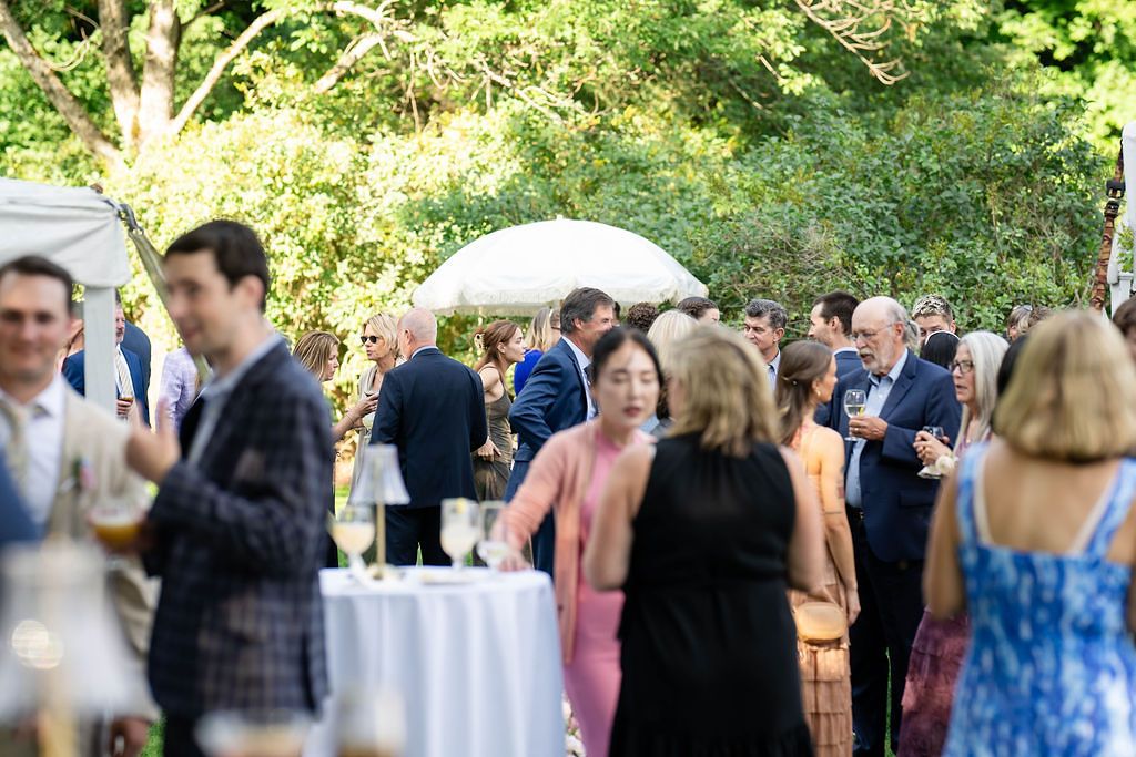 Elegantly dressed wedding guests socializing outdoors near white cocktail tables surrounded by lush greenery.