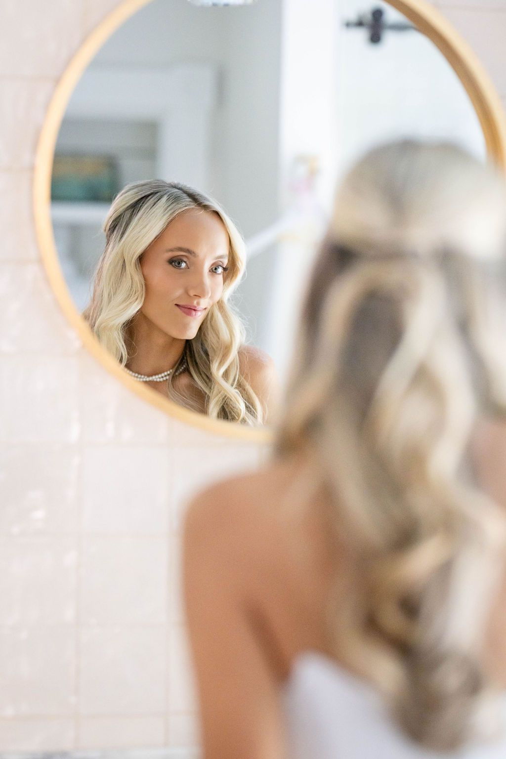 Blonde bride admiring her elegant half-up hairstyle and necklace in a round gold-framed bathroom mirror.