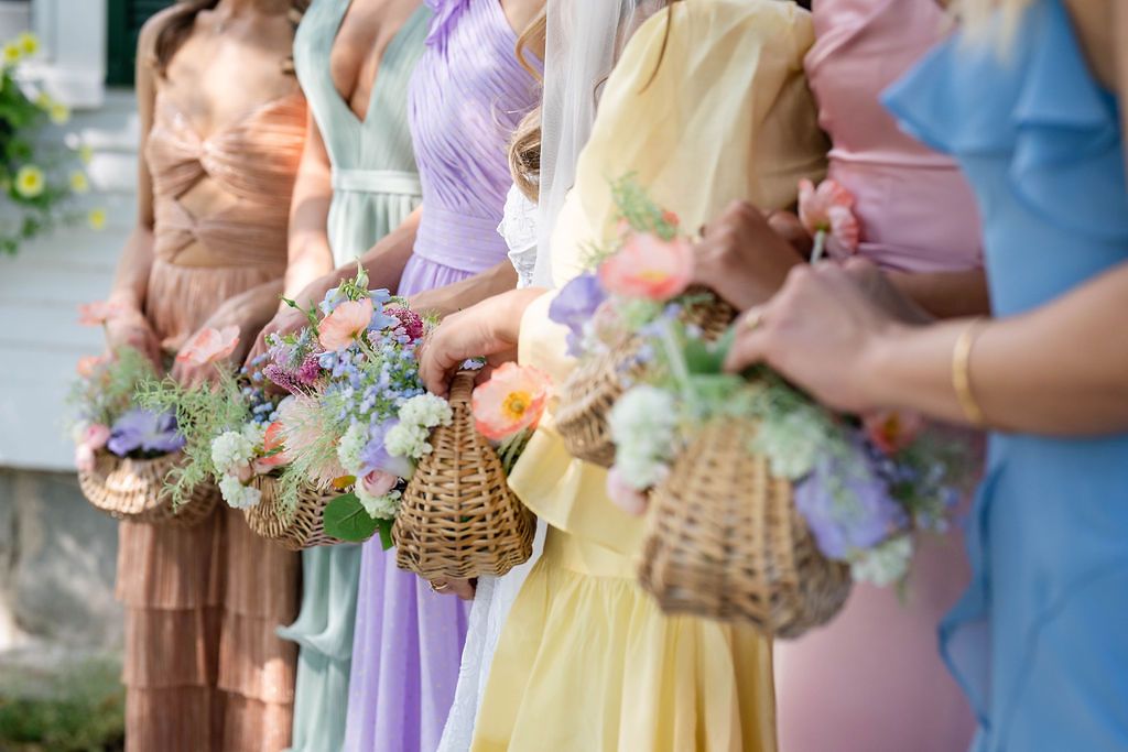 Bridesmaids in pastel mismatched gowns hold wicker flower baskets filled with colorful blooms outdoors.