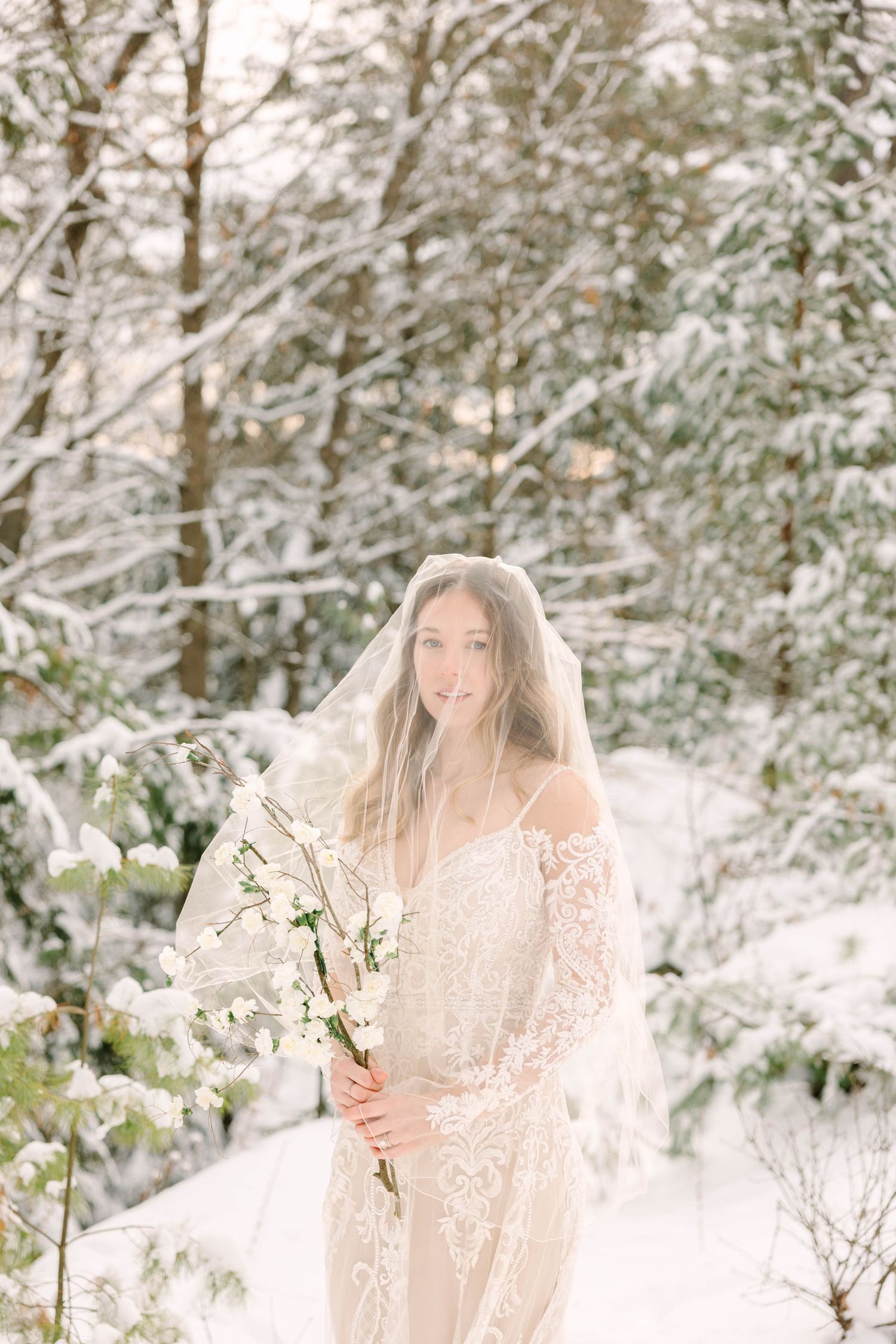 Smiling bride in lace gown and veil holds white floral branches while standing in a bright snowy woodland.
