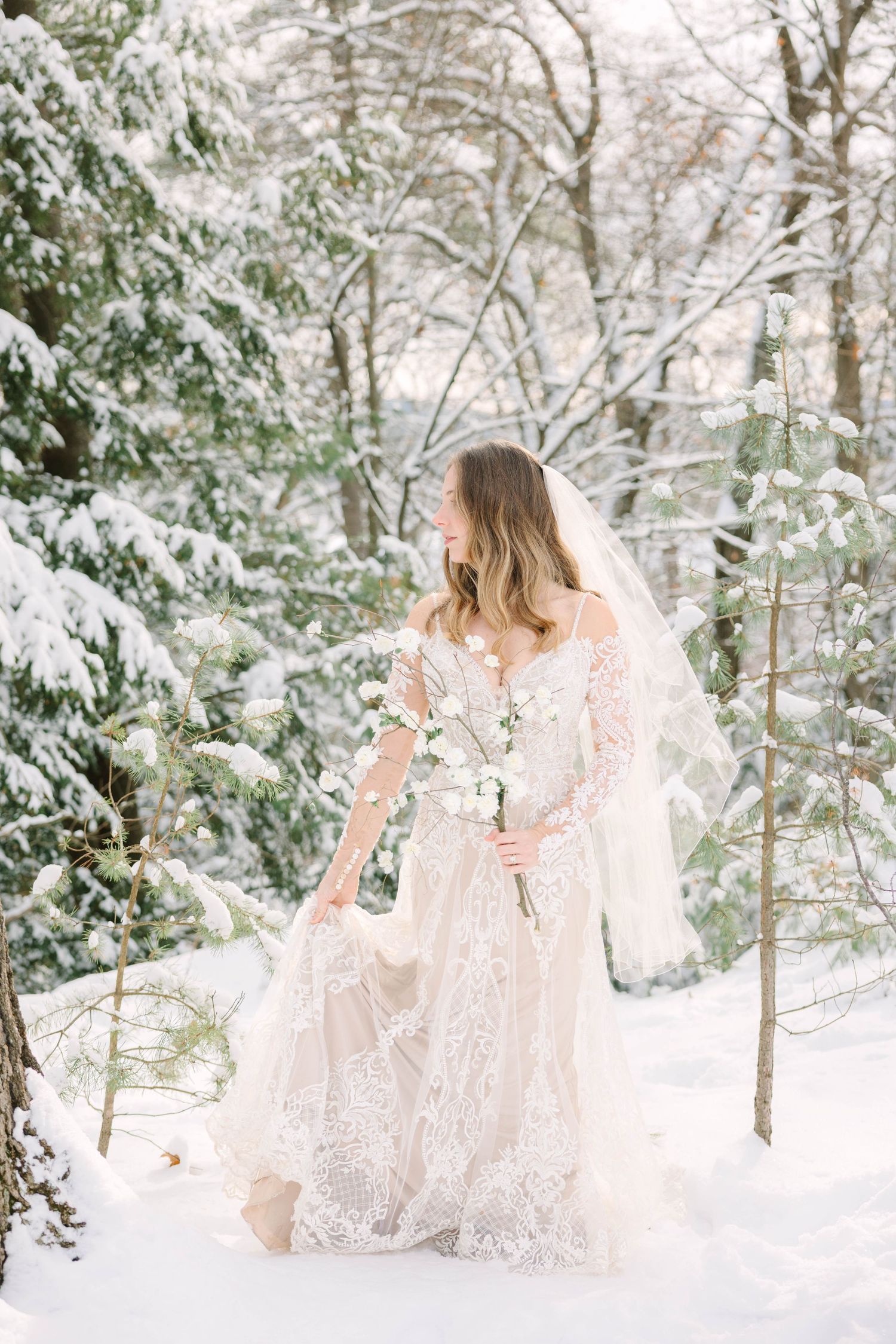 Bride in lace gown and veil holding white bouquet in snowy winter forest wedding photo.