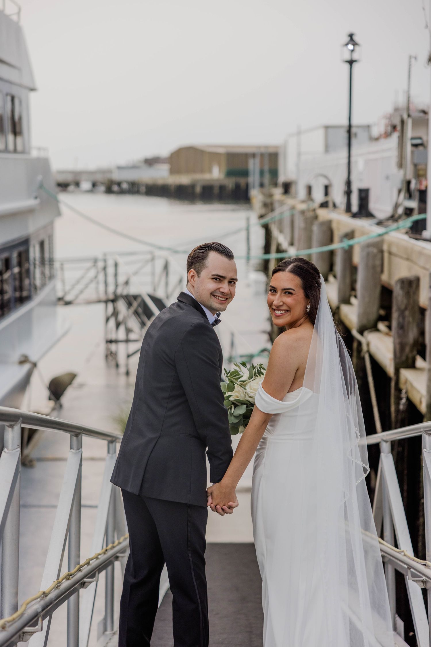 Bride and groom holding hands smiling at camera on waterfront dock near yacht after wedding.