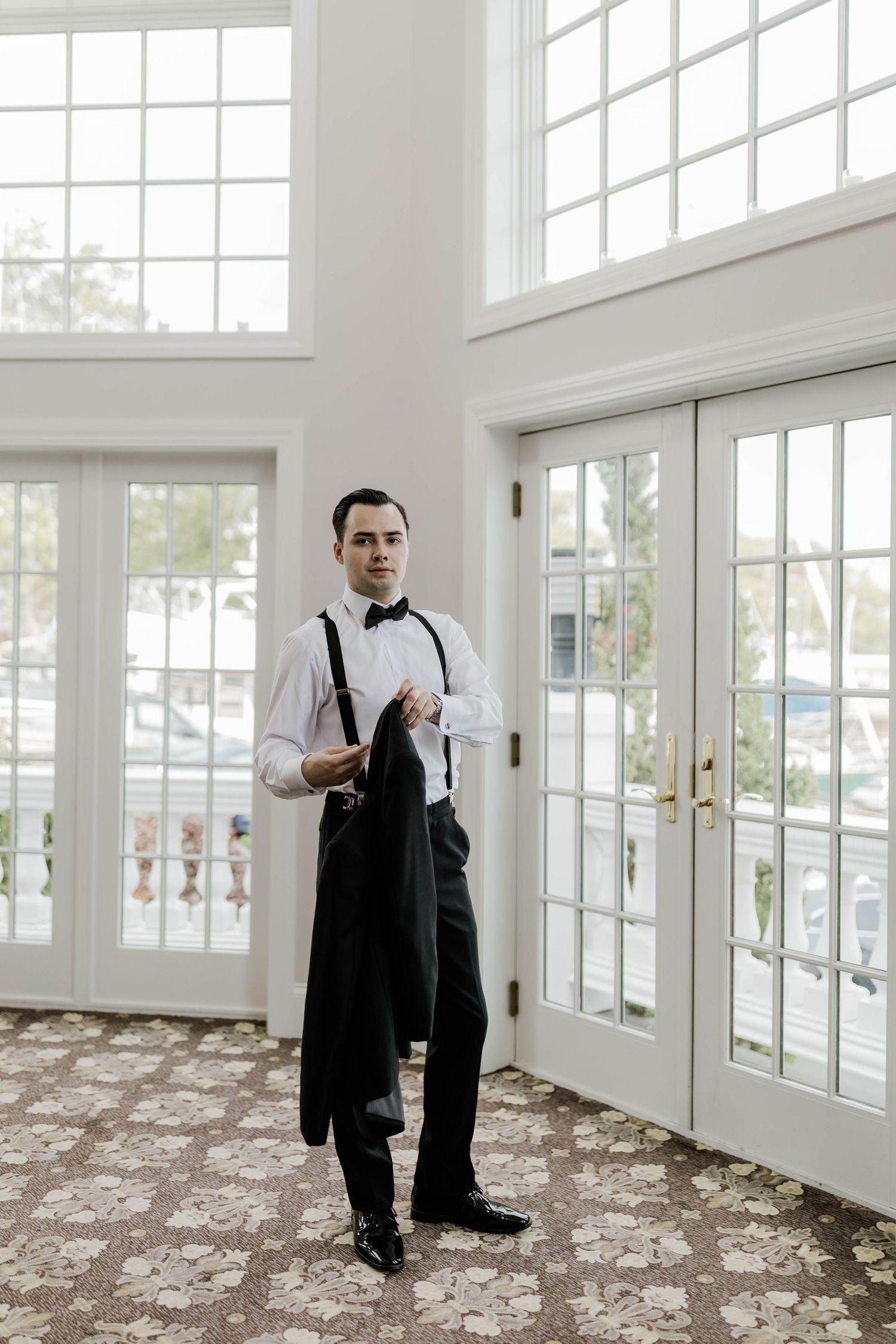 Groom in tuxedo suspenders holding suit jacket standing by tall French doors in elegant venue.