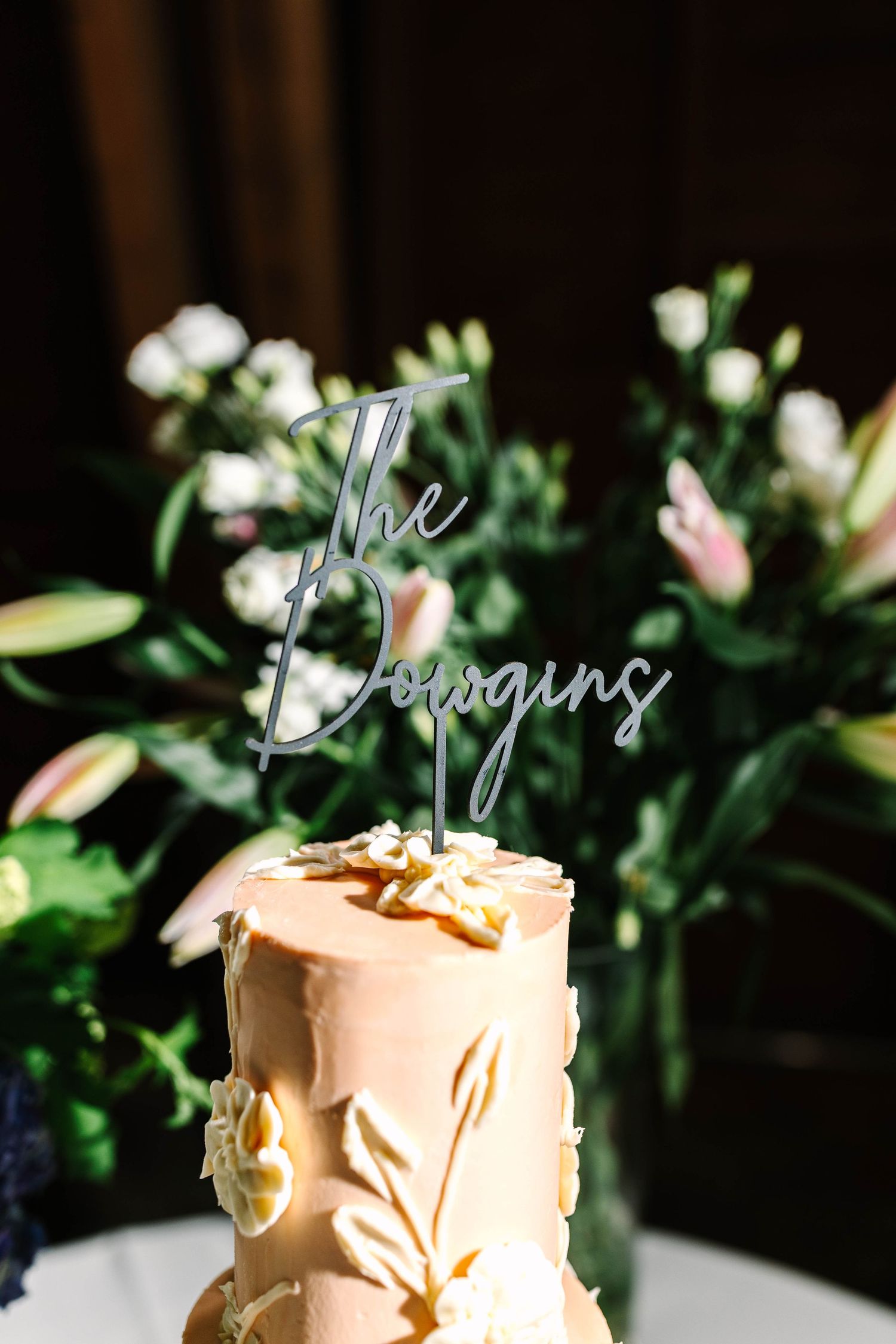 Elegant wedding cake with floral details and The Bowgins cake topper, surrounded by pink and white flowers.