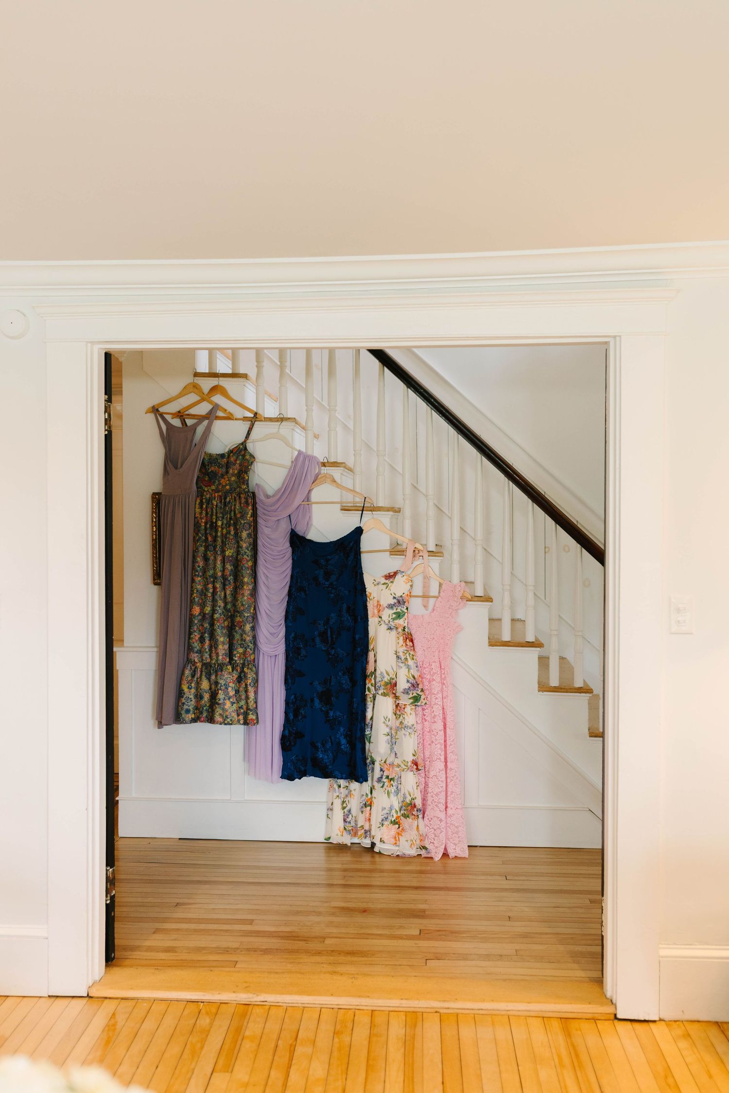 Four bridesmaid dresses in purple, blue, floral, and pink hanging on wooden hangers near a staircase.