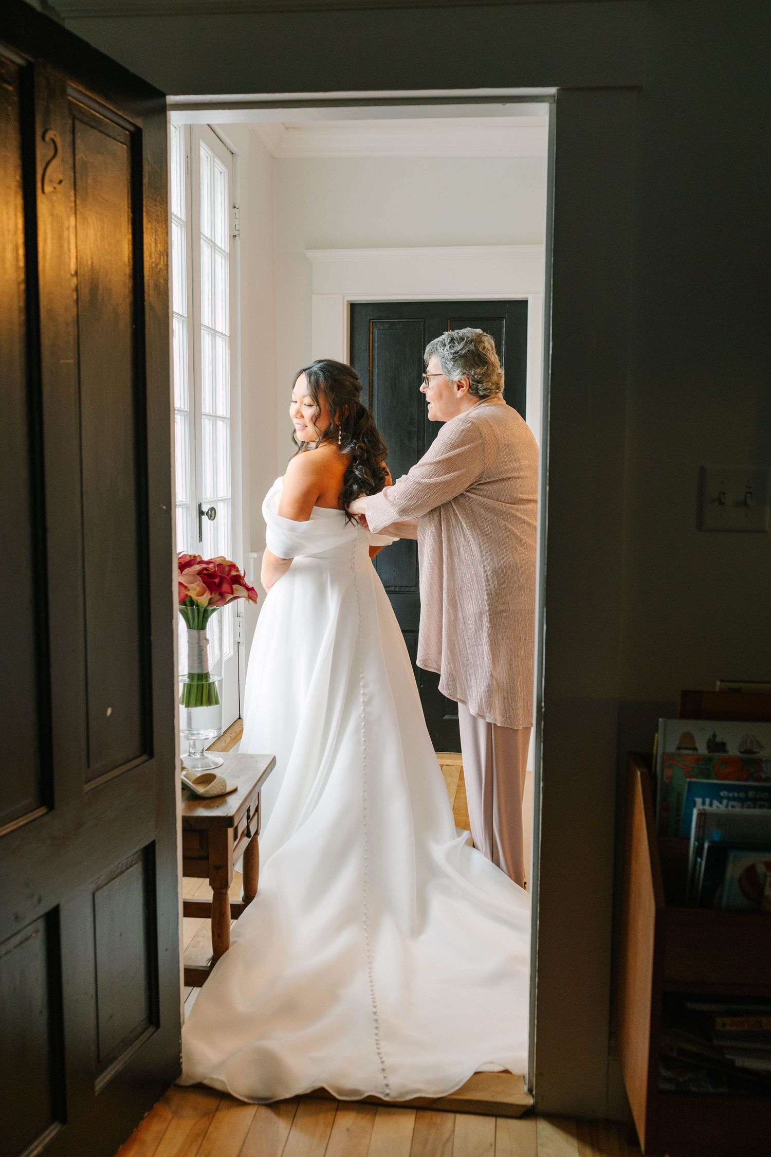 Mother helps bride fasten her white wedding gown in a bright doorway before the ceremony.