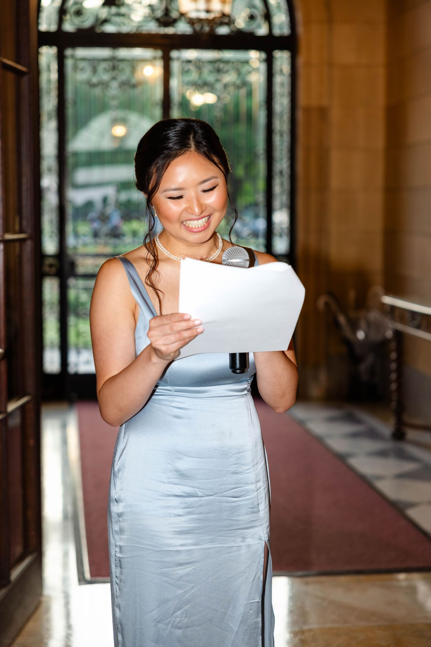 Smiling bridesmaid in silver satin gown reads speech paper at elegant wedding venue.