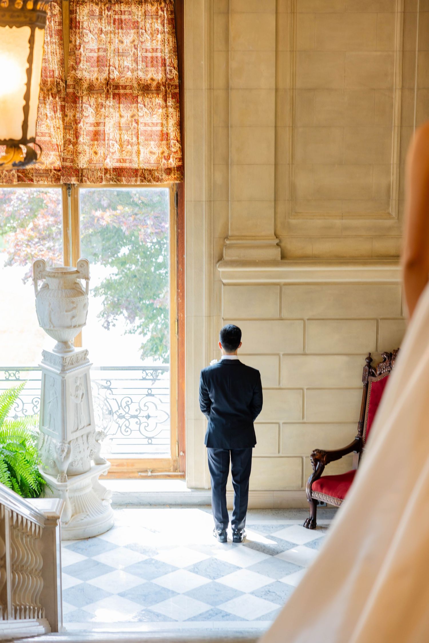 Groom in black tuxedo stands waiting by grand window in marble-columned hall before first look reveal.