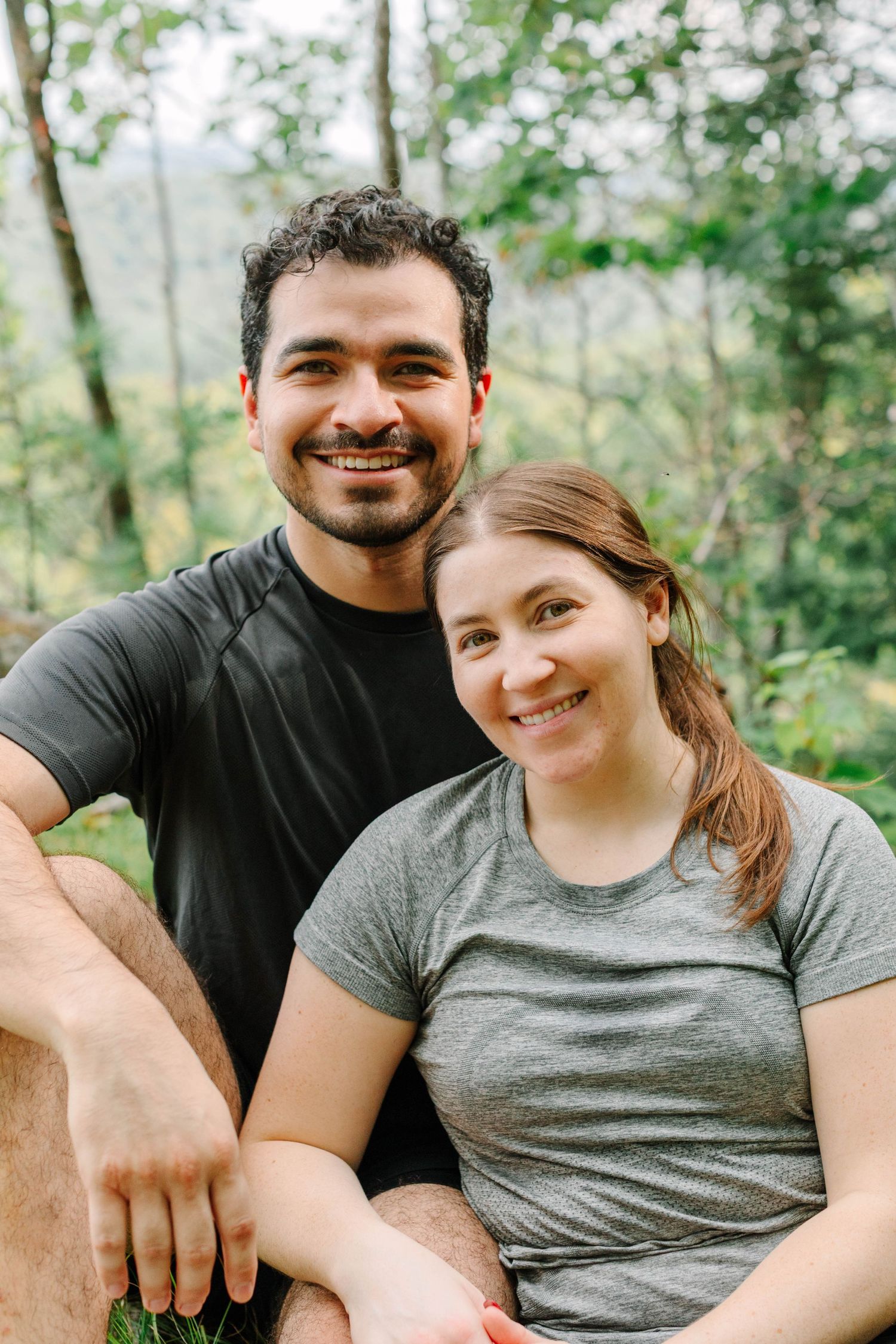 Smiling couple sitting together outdoors in a lush green forest setting, wearing casual athletic wear.