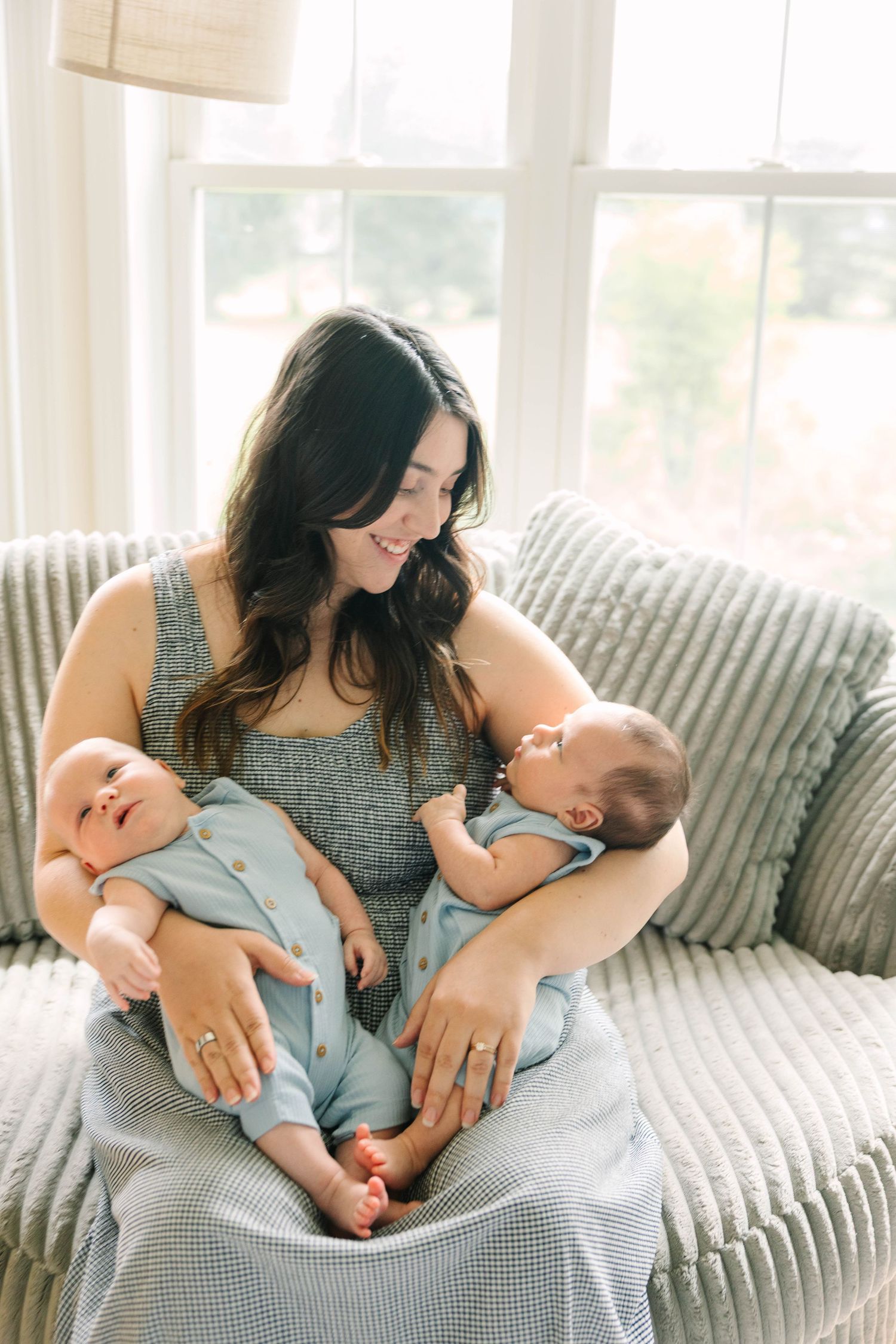 Brunette mother in blue dress smiling at her twin newborns while seated on a gray sofa.