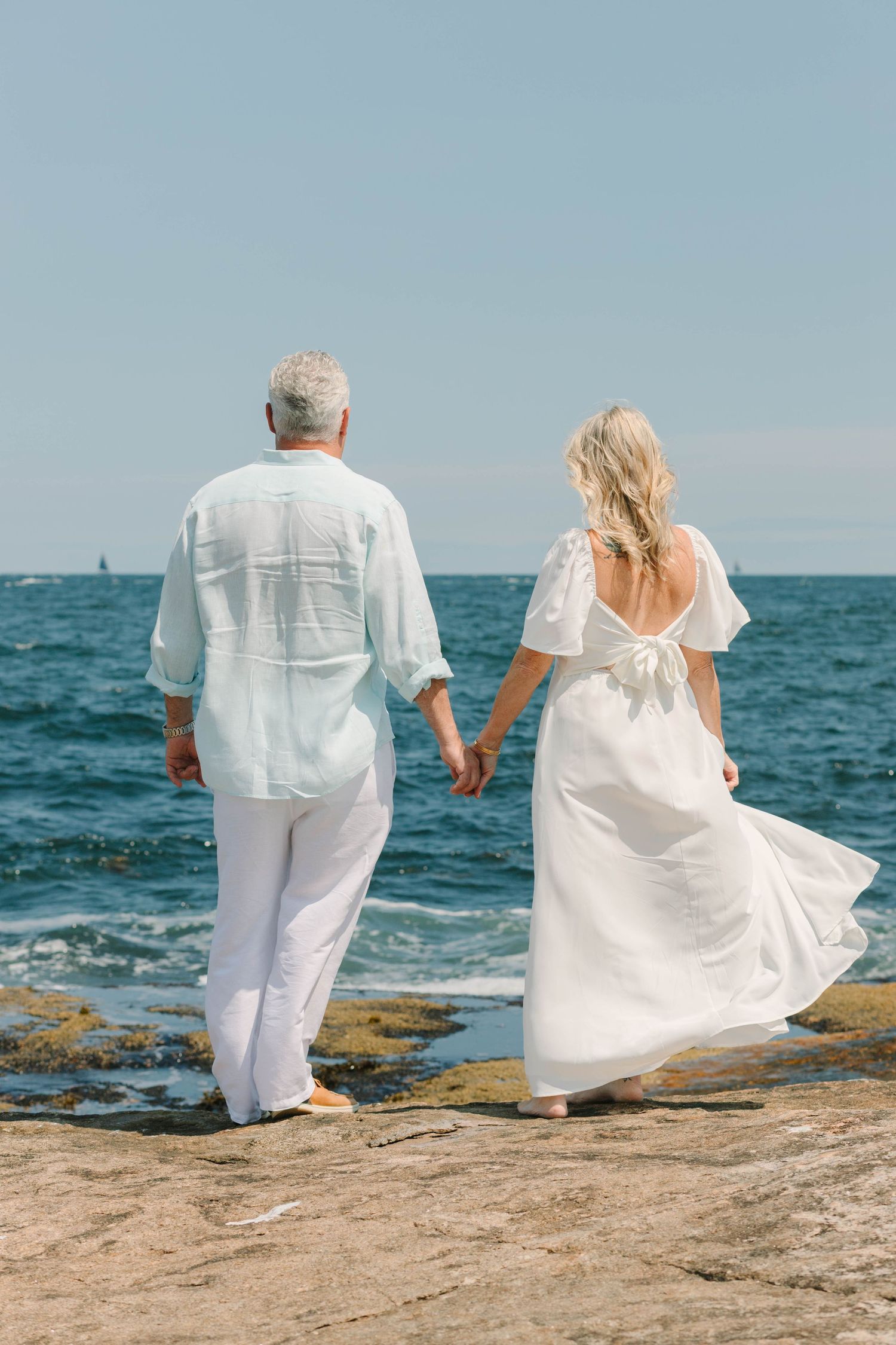 Couple in white outfits holding hands on rocky shore by the ocean on a sunny day.
