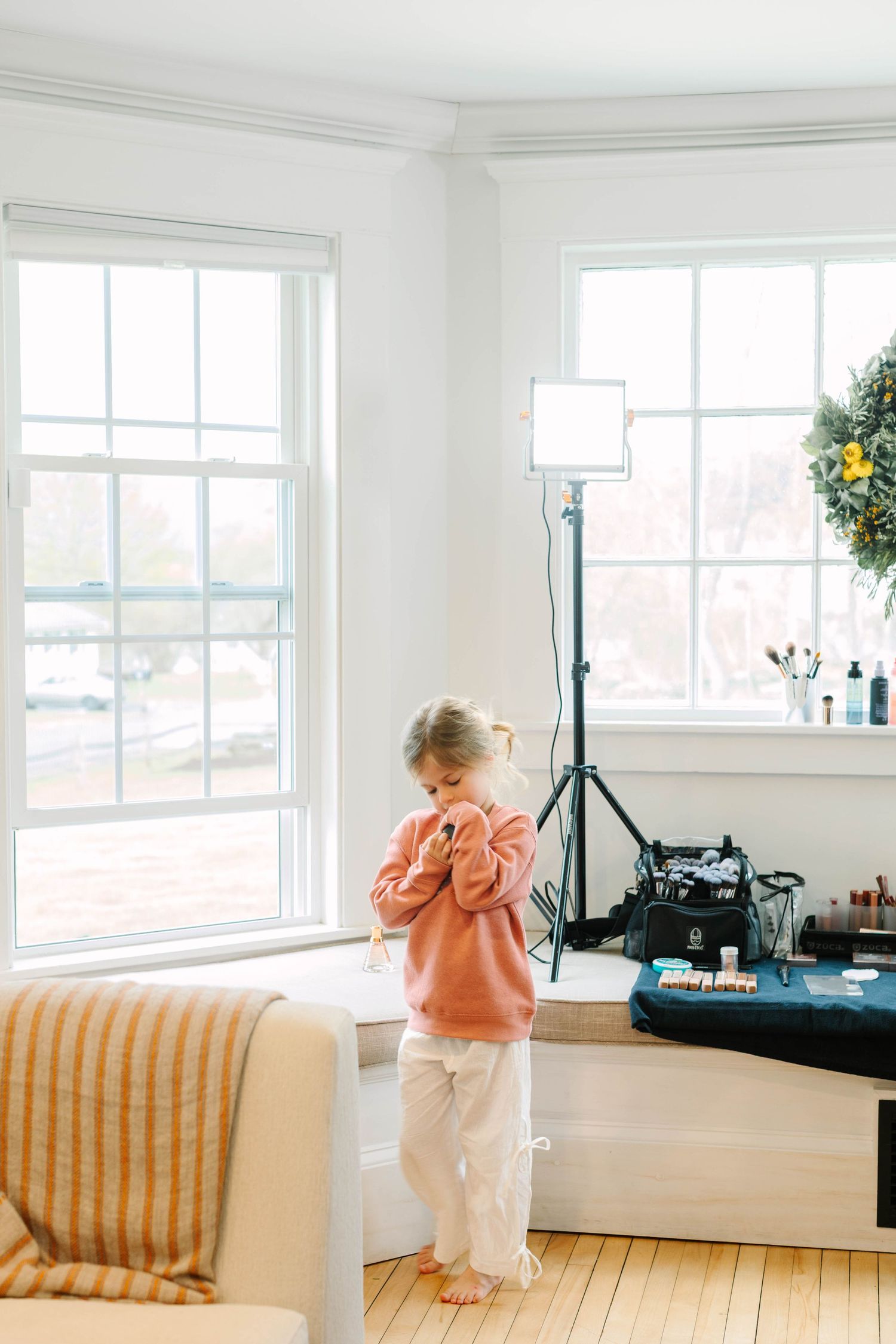 Young girl in pink sweatshirt stands by bright windows near photography lighting equipment in a white room.