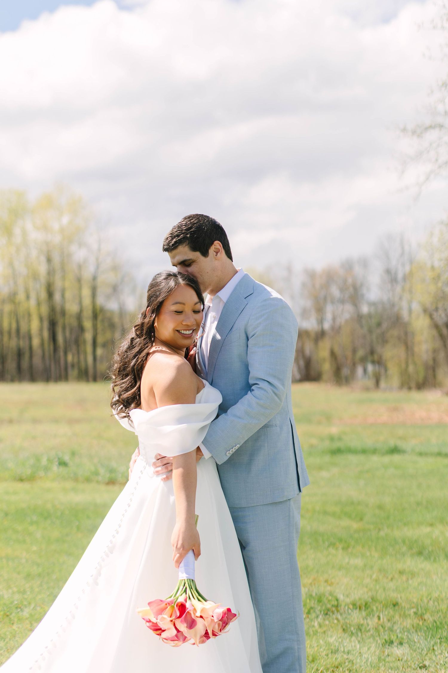 Bride and groom embracing in a green field, bride holding pink tulip bouquet on wedding day.