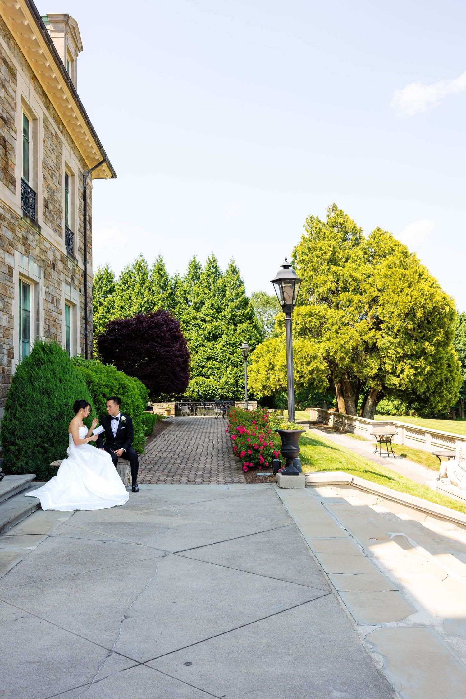 Bride and groom sit together outside a stone mansion surrounded by lush greenery on wedding day.