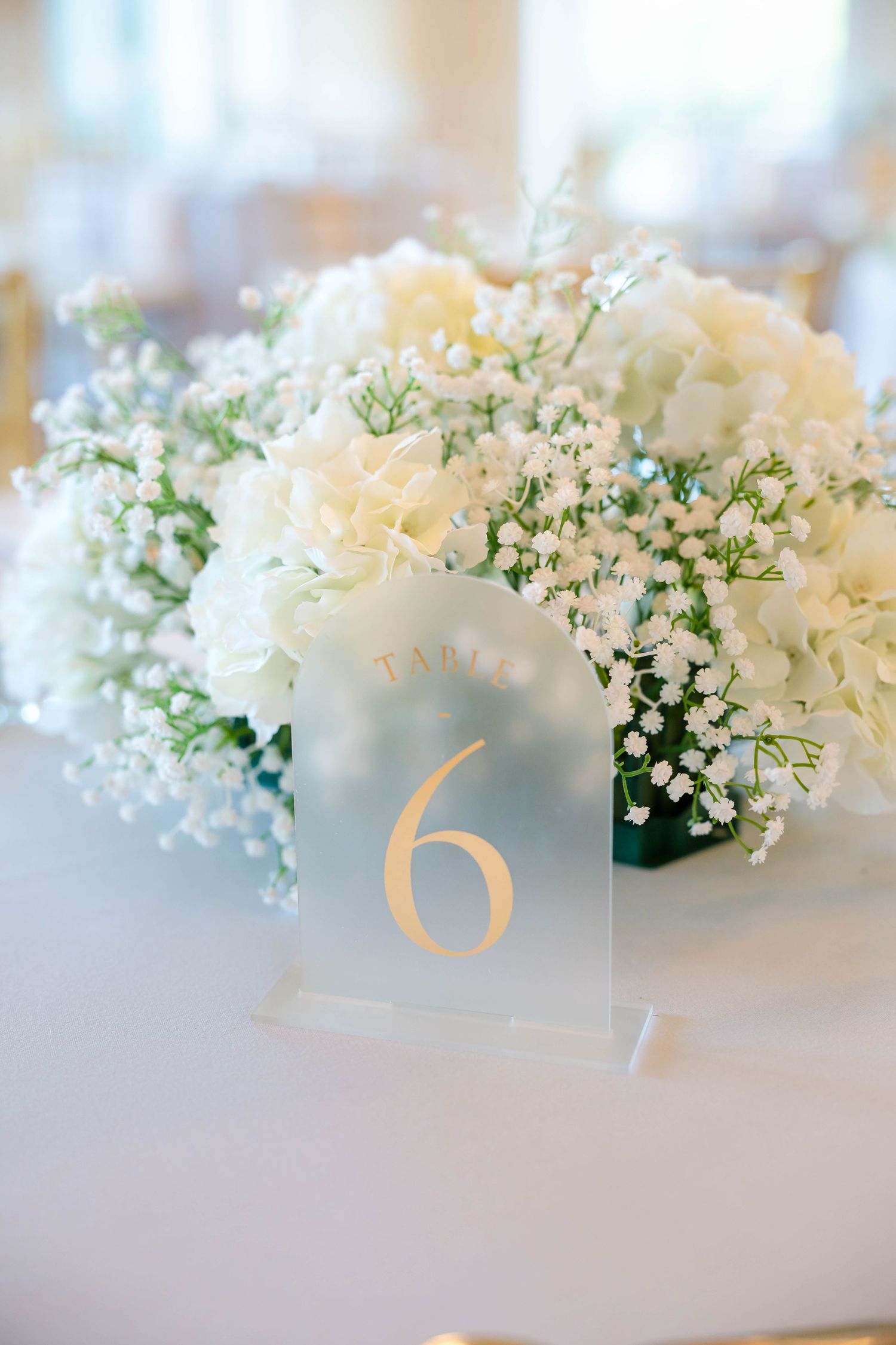 Elegant white hydrangea and baby's breath centerpiece with gold table number six sign.