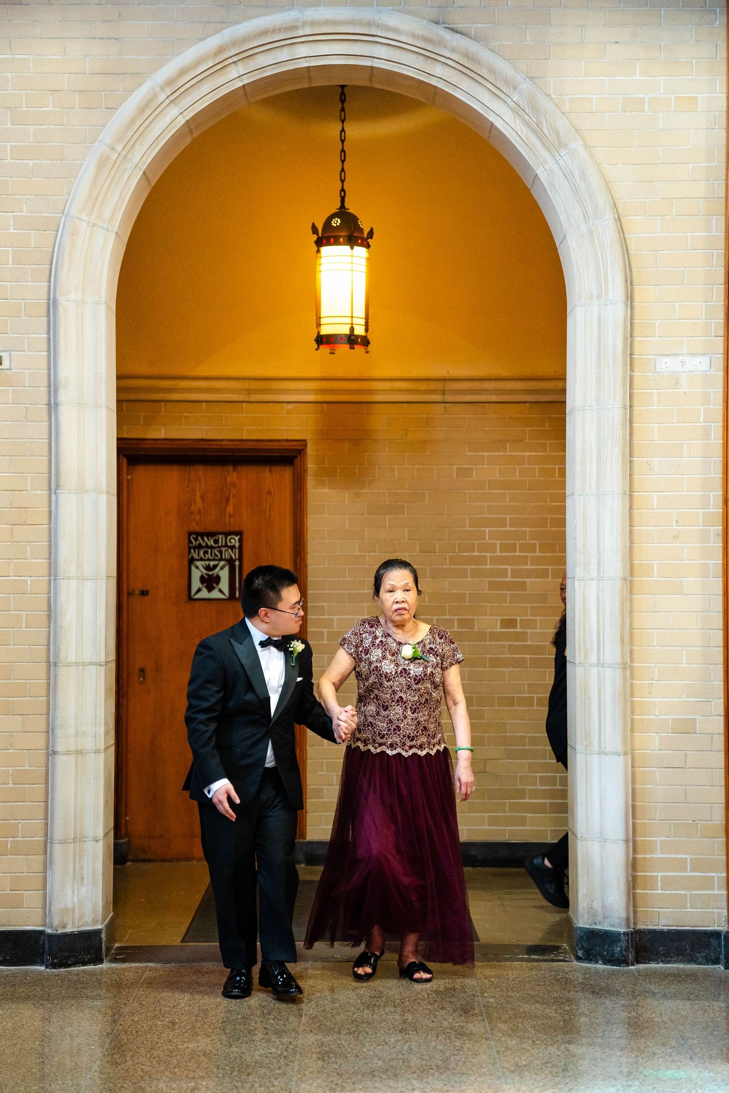 Groom in tuxedo escorts mother in burgundy gown through stone archway of a classic church venue.