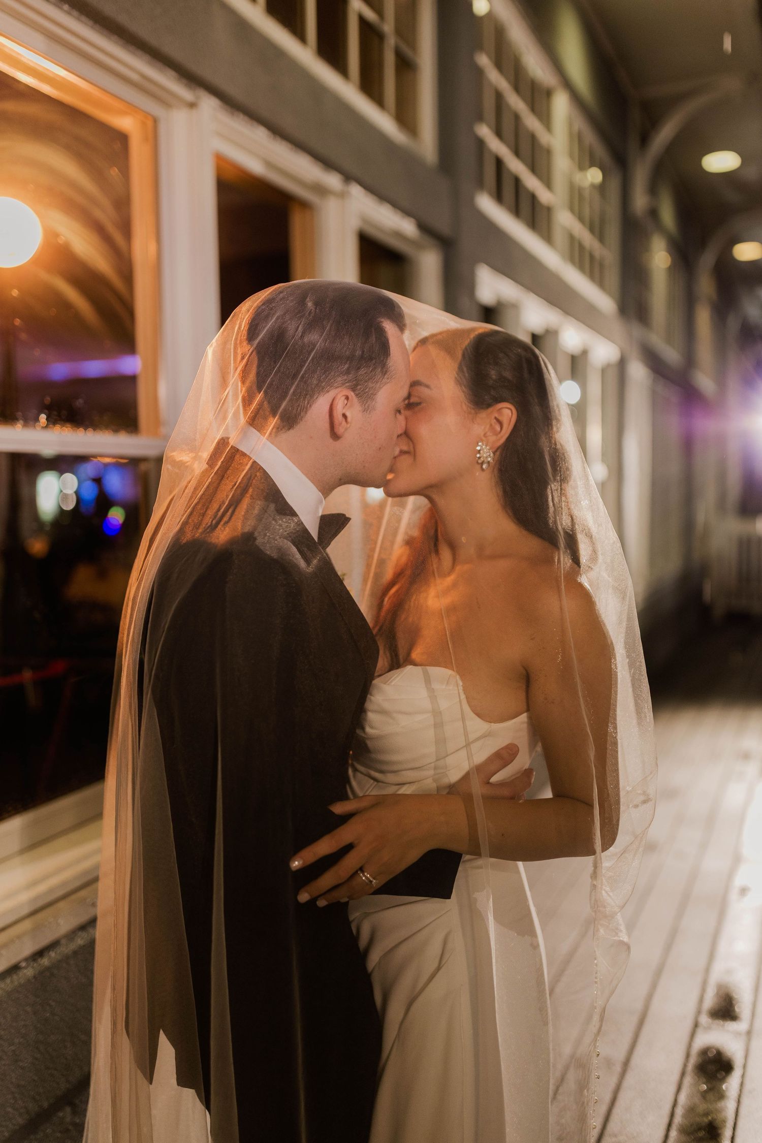 Bride and groom share romantic kiss under flowing veil outside at night with warm glowing lights behind them.