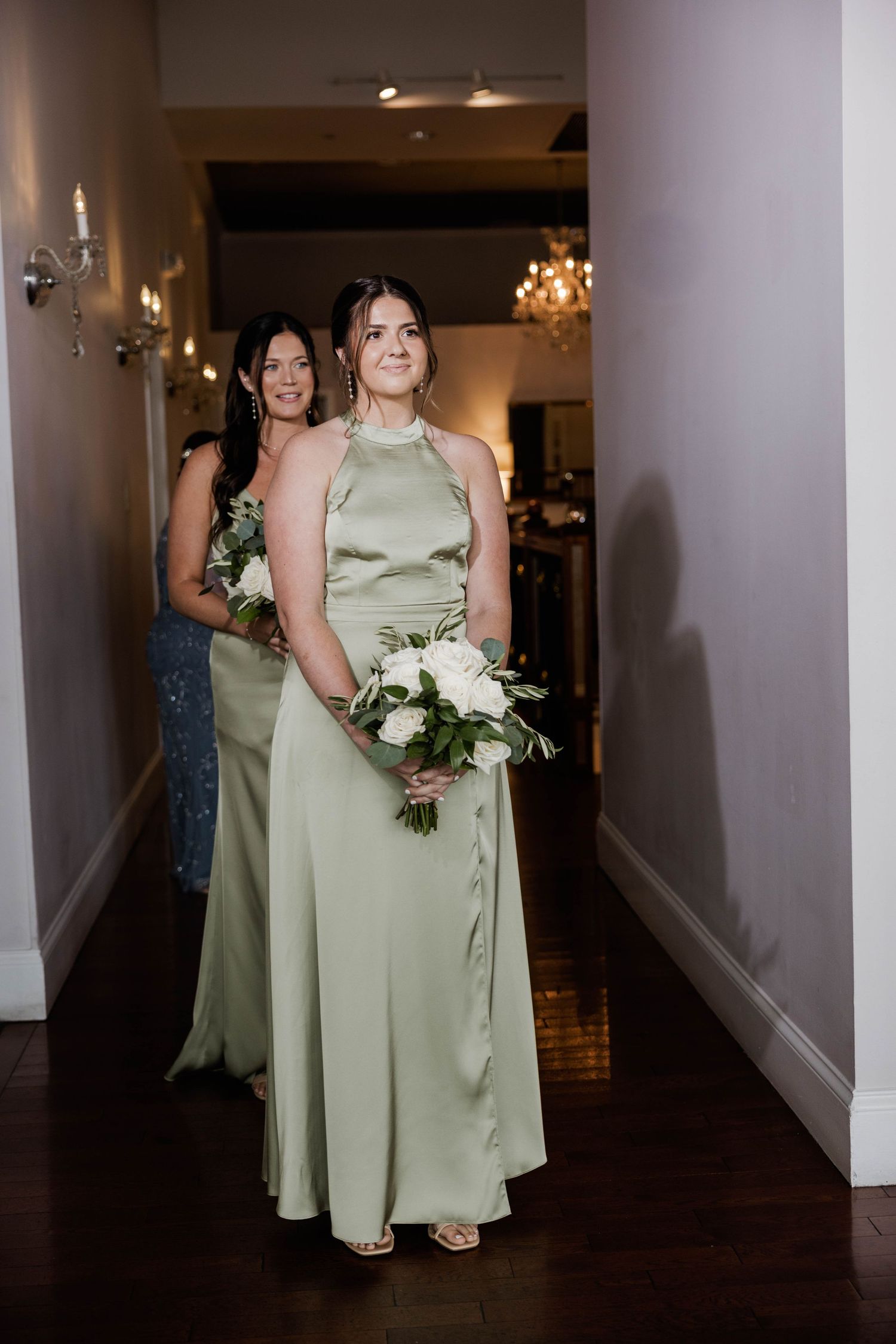 Bridesmaid in sage green satin halter gown carrying white floral bouquet walking down hallway.