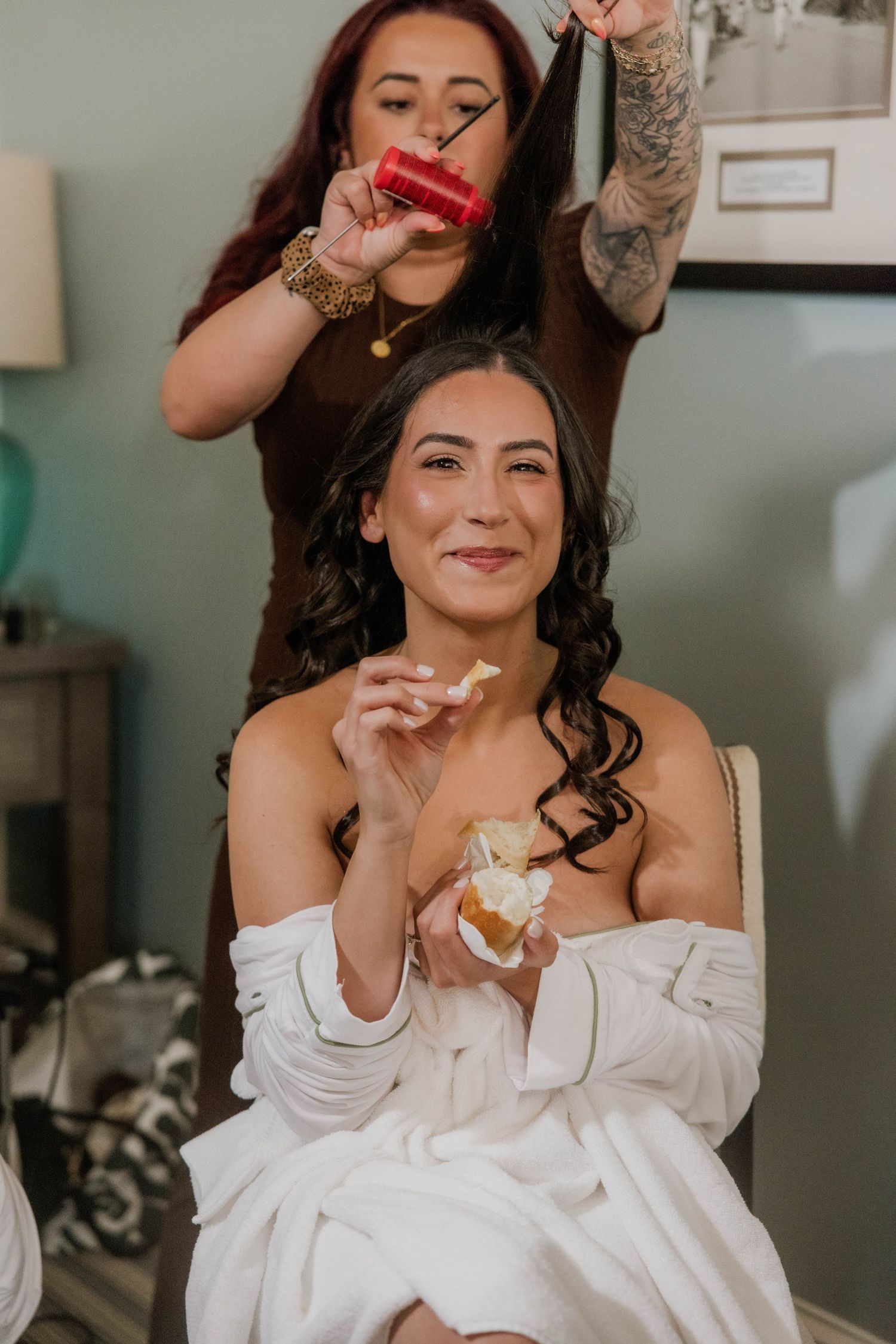 Bride getting hair done while eating a snack in a white robe before her wedding ceremony.